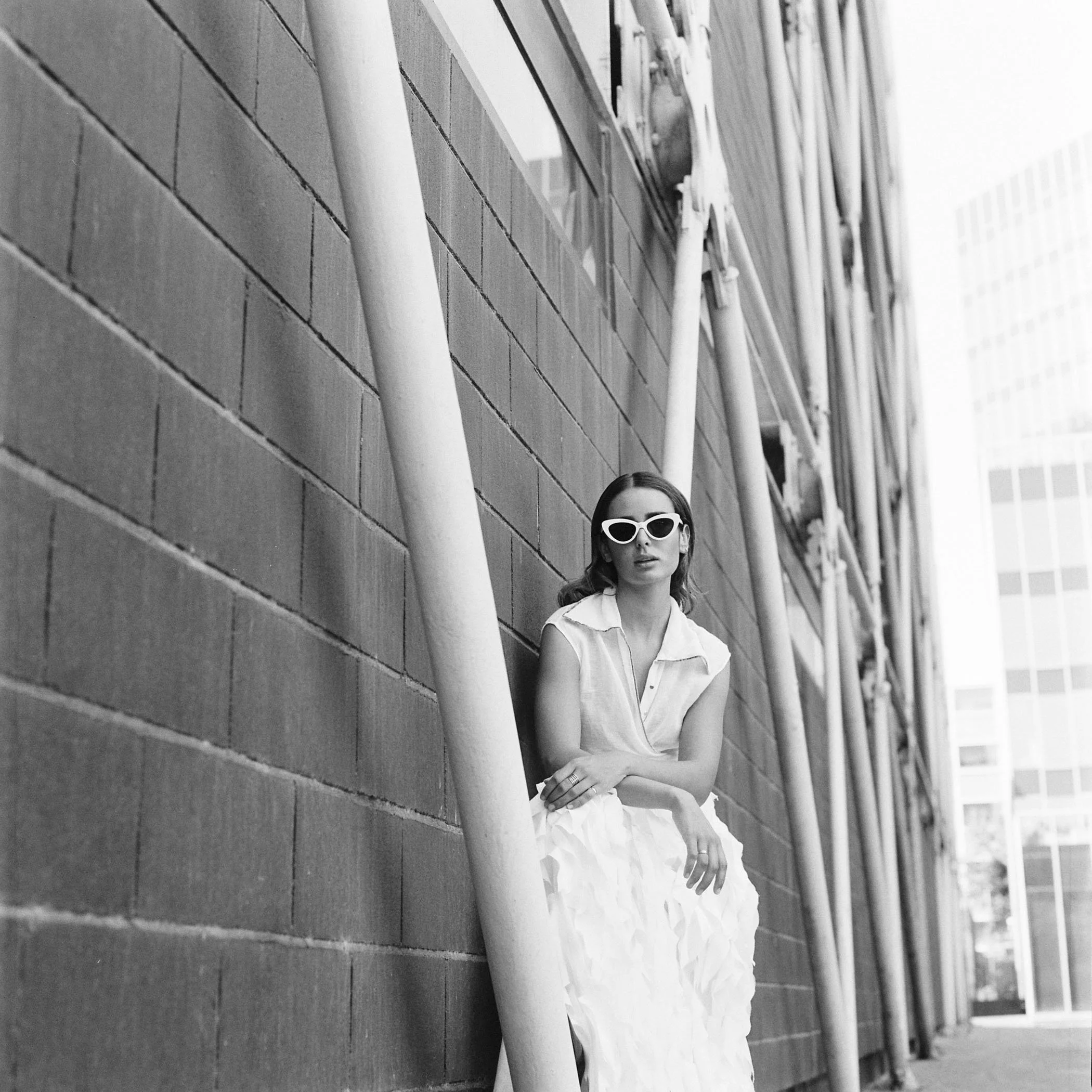 Mujer con gafas blancas sentada junto a una pared de ladrillos y estructuras metálicas en un entorno urbano.
