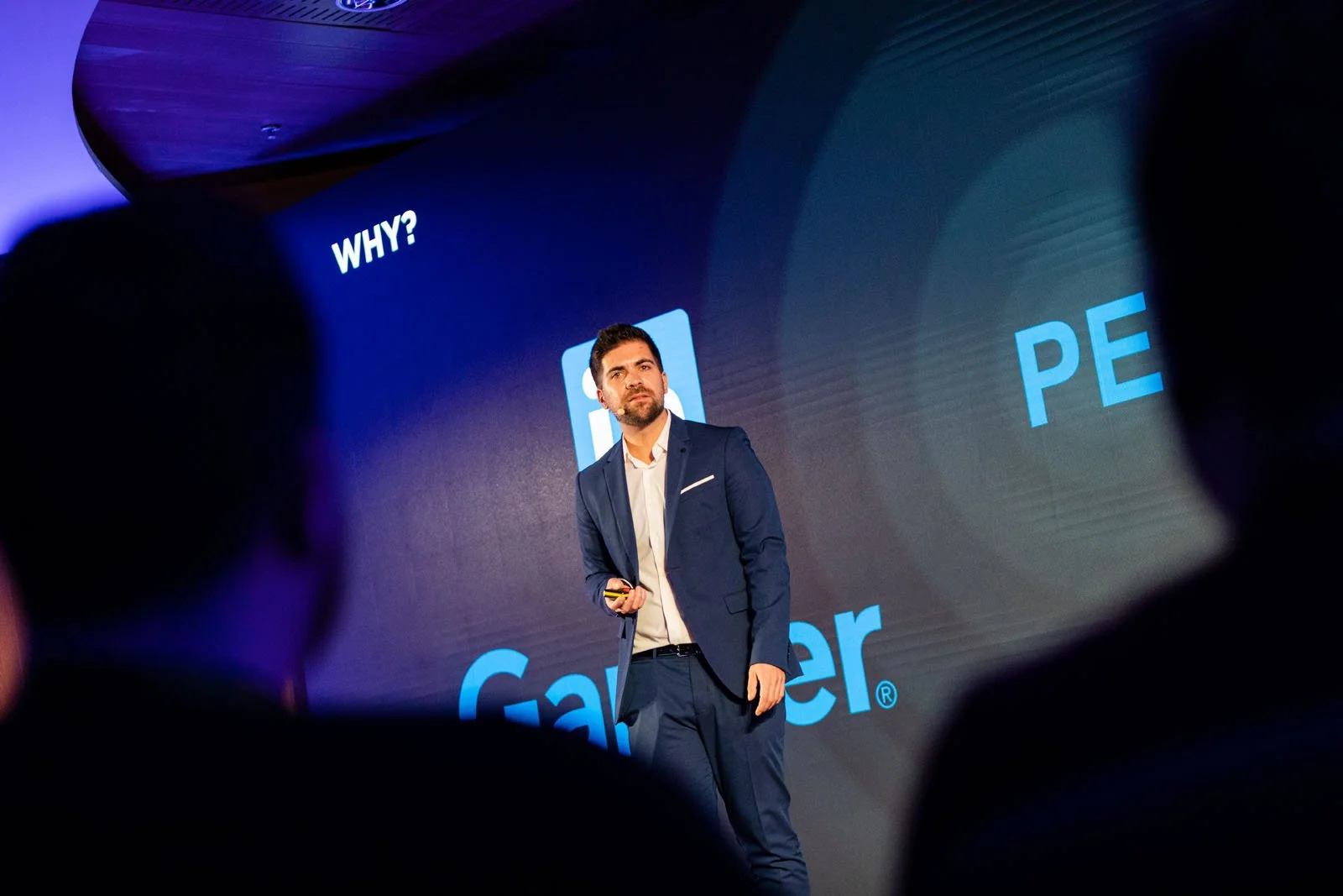 Hombre de negocios con barba y vestido con traje azul, presentando en un escenario con una pantalla grande que muestra el logo de Google y las palabras en inglés 'WHY?' y parcialmente 'PER'.