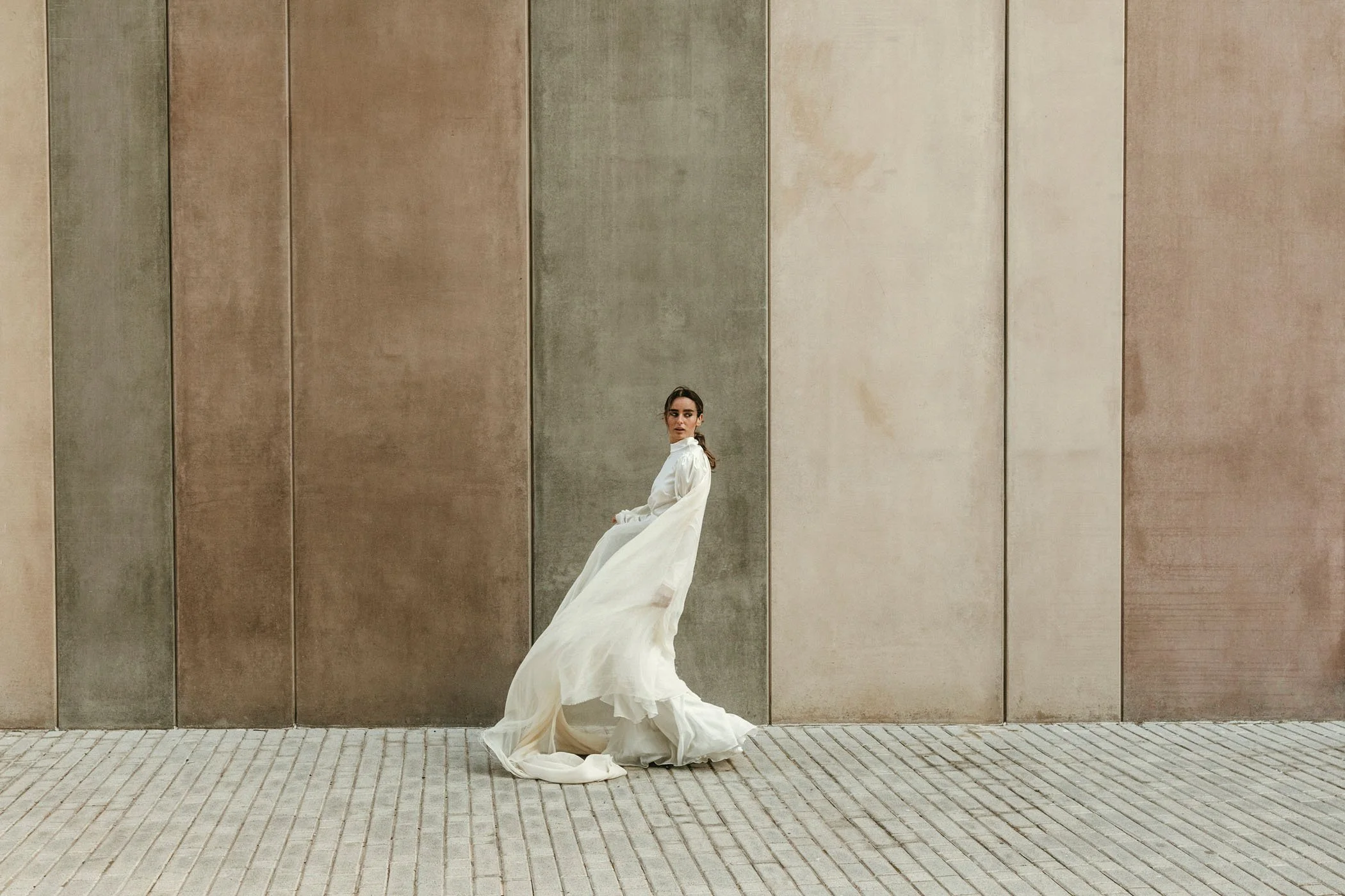 Mujer vestida con vestido blanco y capa, sentada contra una pared de colores neutros en un espacio abierto.