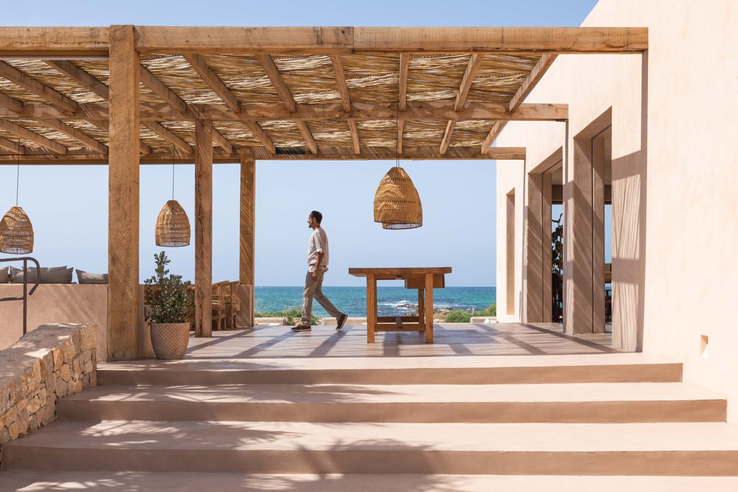 Hombre caminando por una terraza con vista al mar, estructura de madera y decoración de estilo costero.
