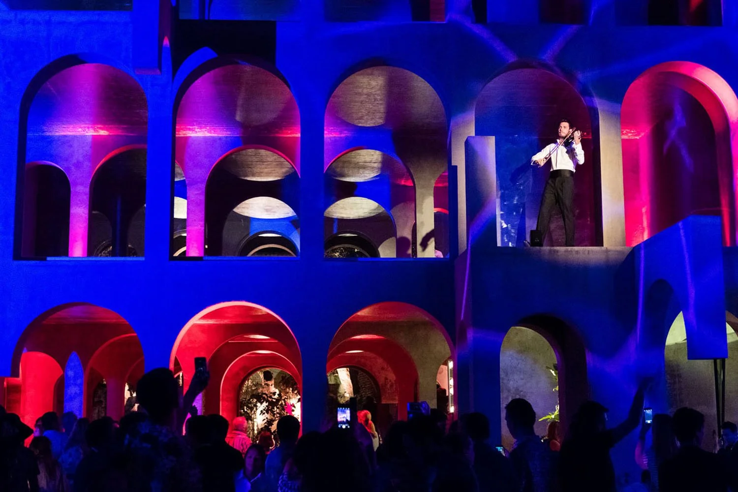 Un concierto con un artista tocando violín en un escenario de estructura moderna con arcos iluminados en colores rosa, morado y azul, con público disfrutando la actuación.