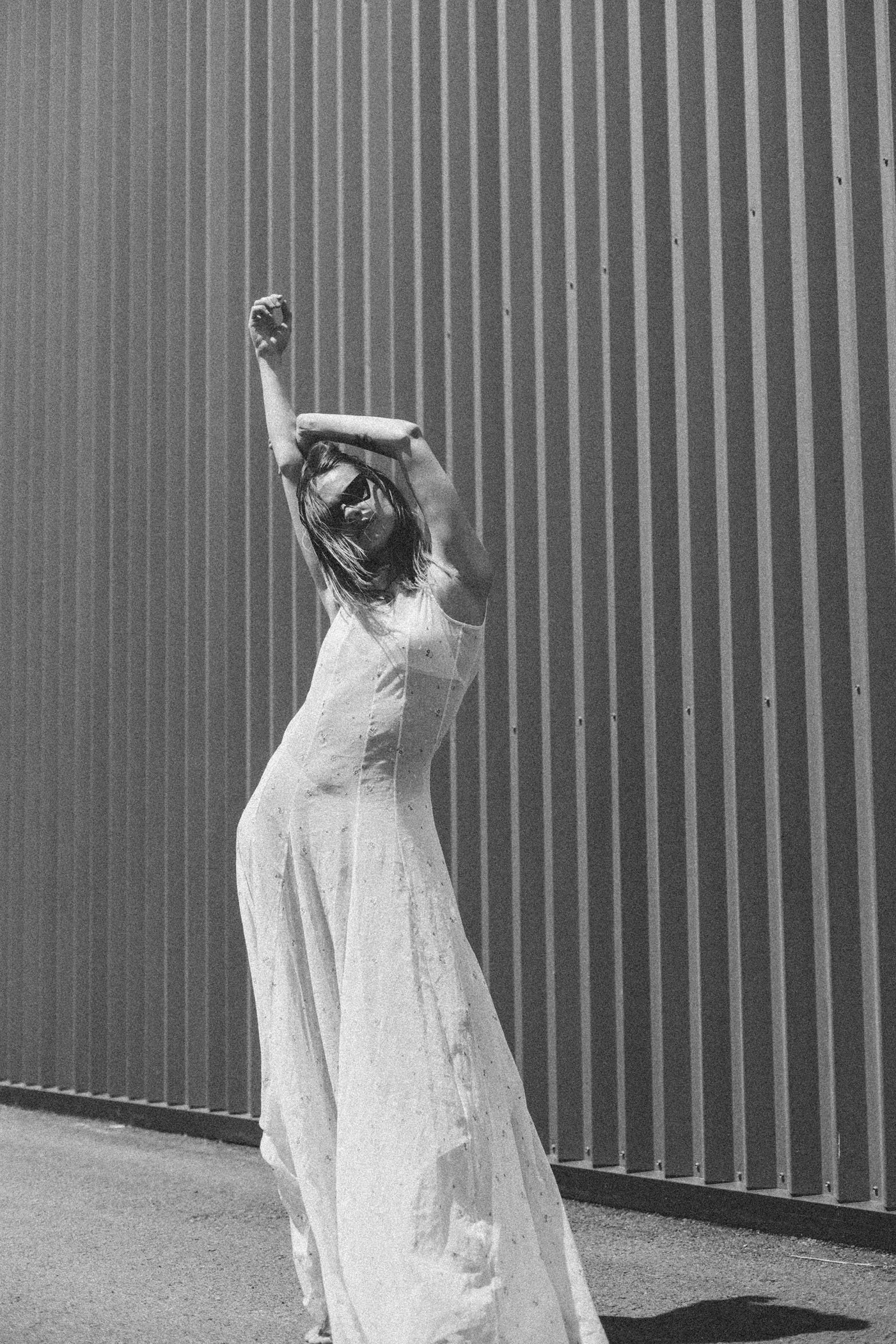 Mujer con vestido largo y gafas de sol posando frente a una pared de listones verticales en una fotografía en blanco y negro.