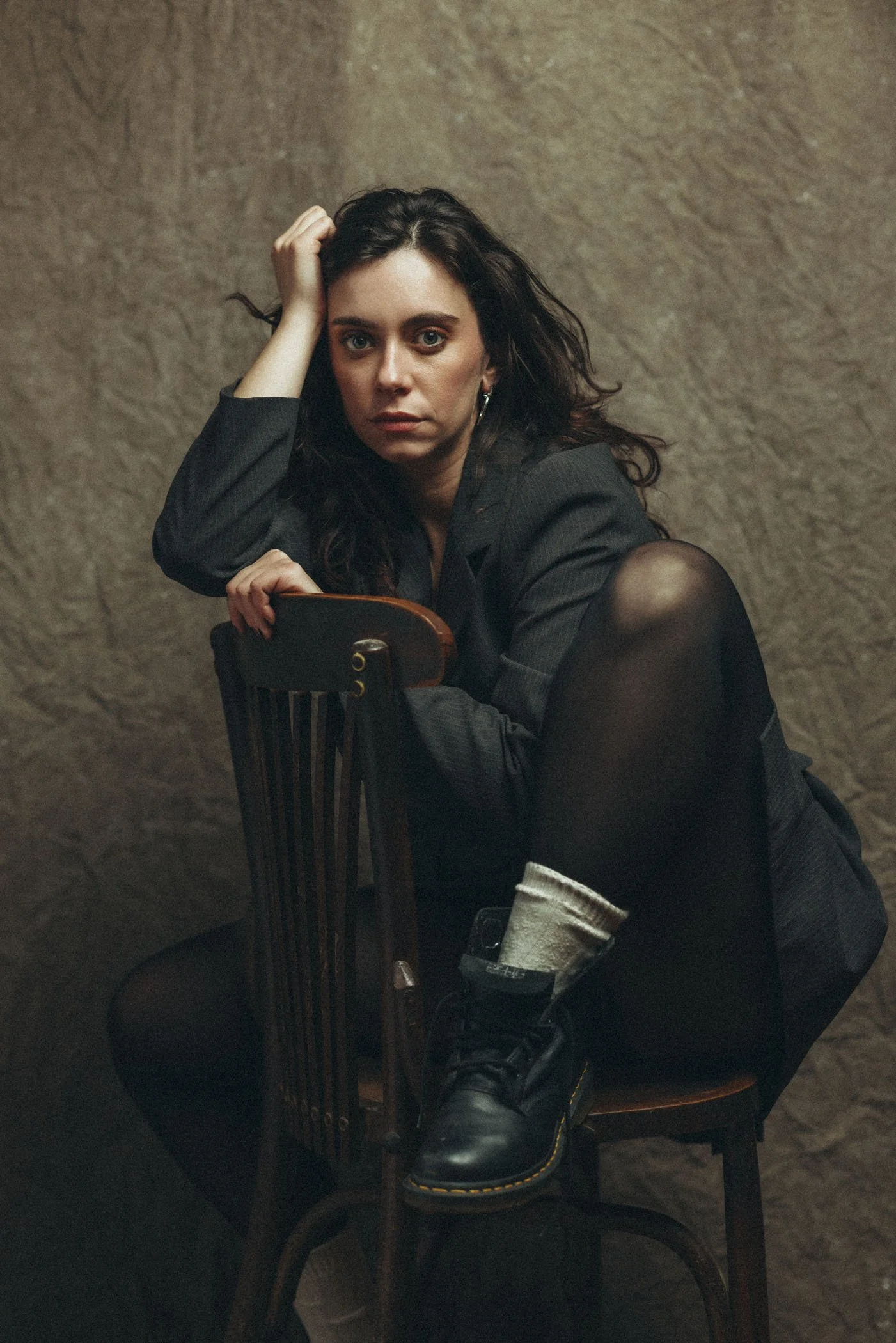 Mujer con cabello rizado oscuro, vista frontal, en pose pensativa, usando ropa oscura, camiseta de manga larga y falda, sentada en una silla de madera, con expresión seria y fondo de pared texturizada.