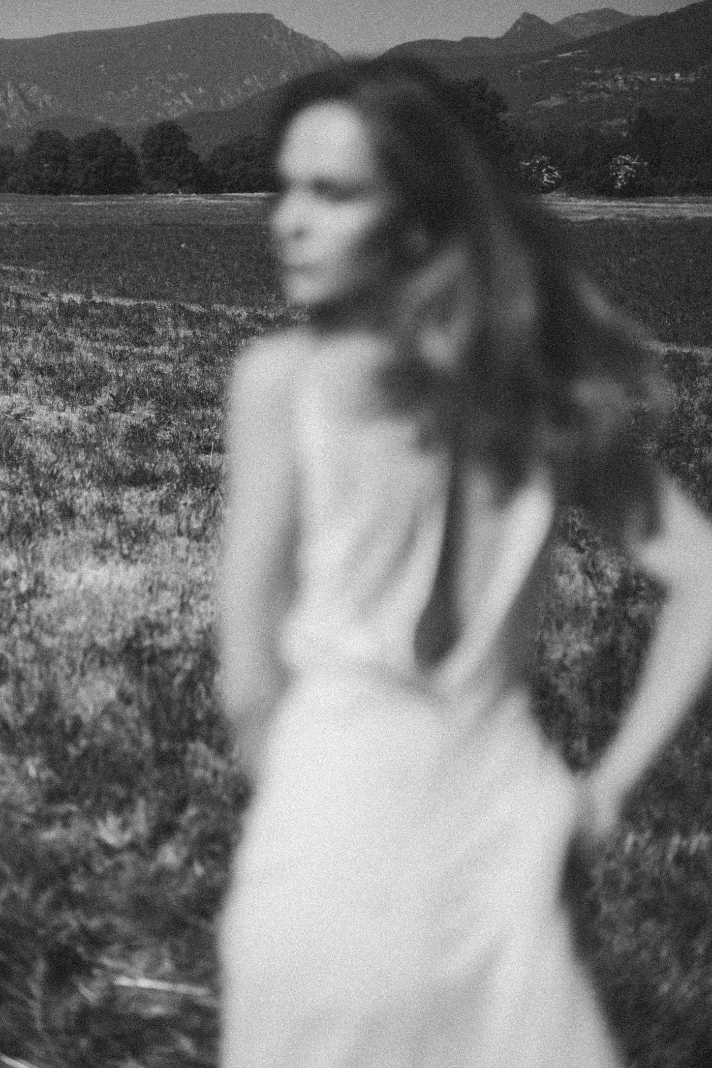 Una mujer de espaldas en un campo, con montañas al fondo, en blanco y negro, vista borrosa.