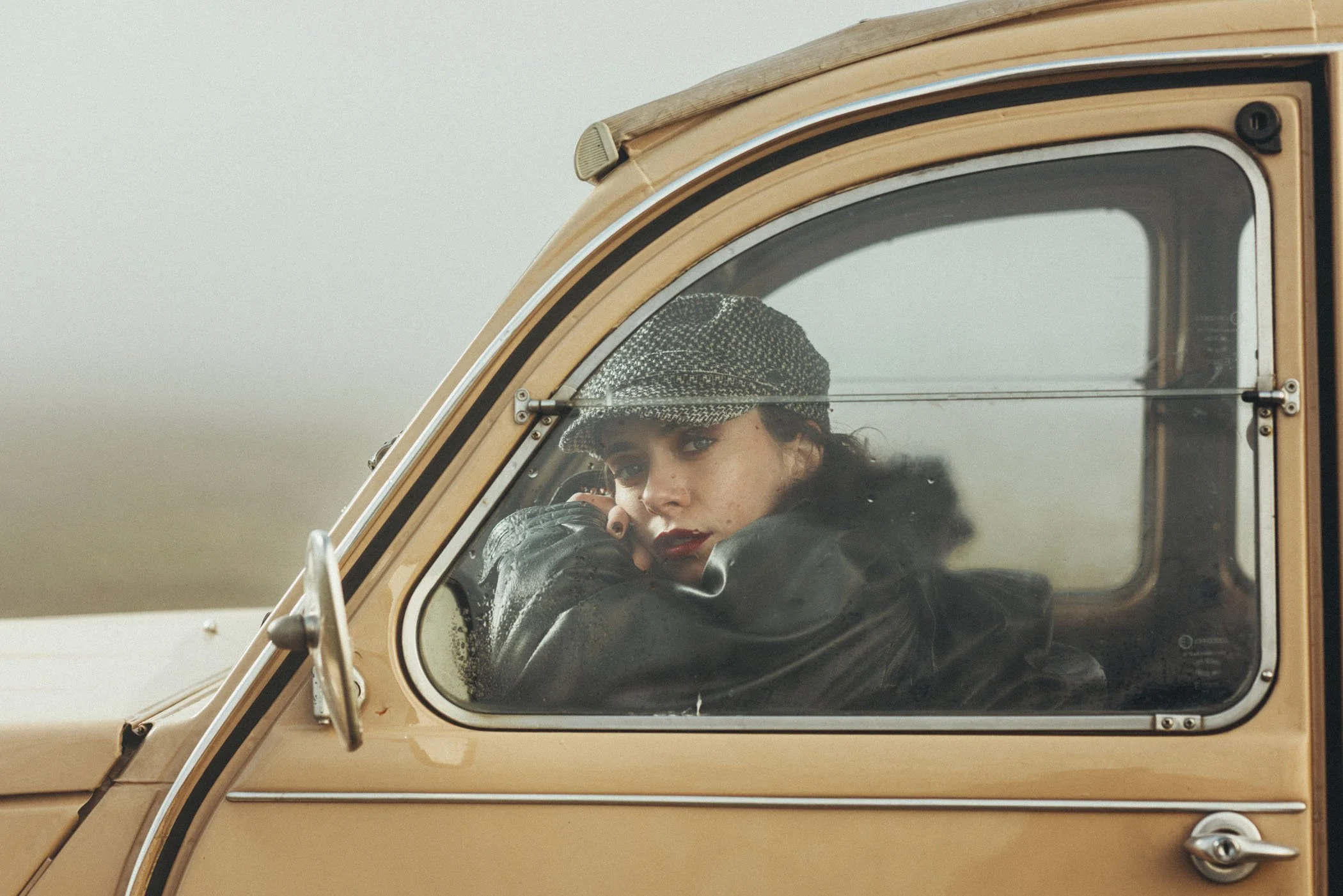 Mujer con gorra y chaqueta negra mira por la ventana de un vehículo clásico de color beige.