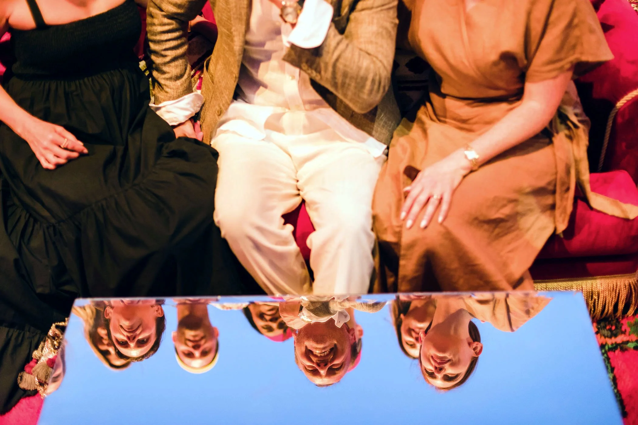 Grupo de personas sentadas en un sofá, sonriendo y conversando, con un espejo que refleja sus rostros y la escena.