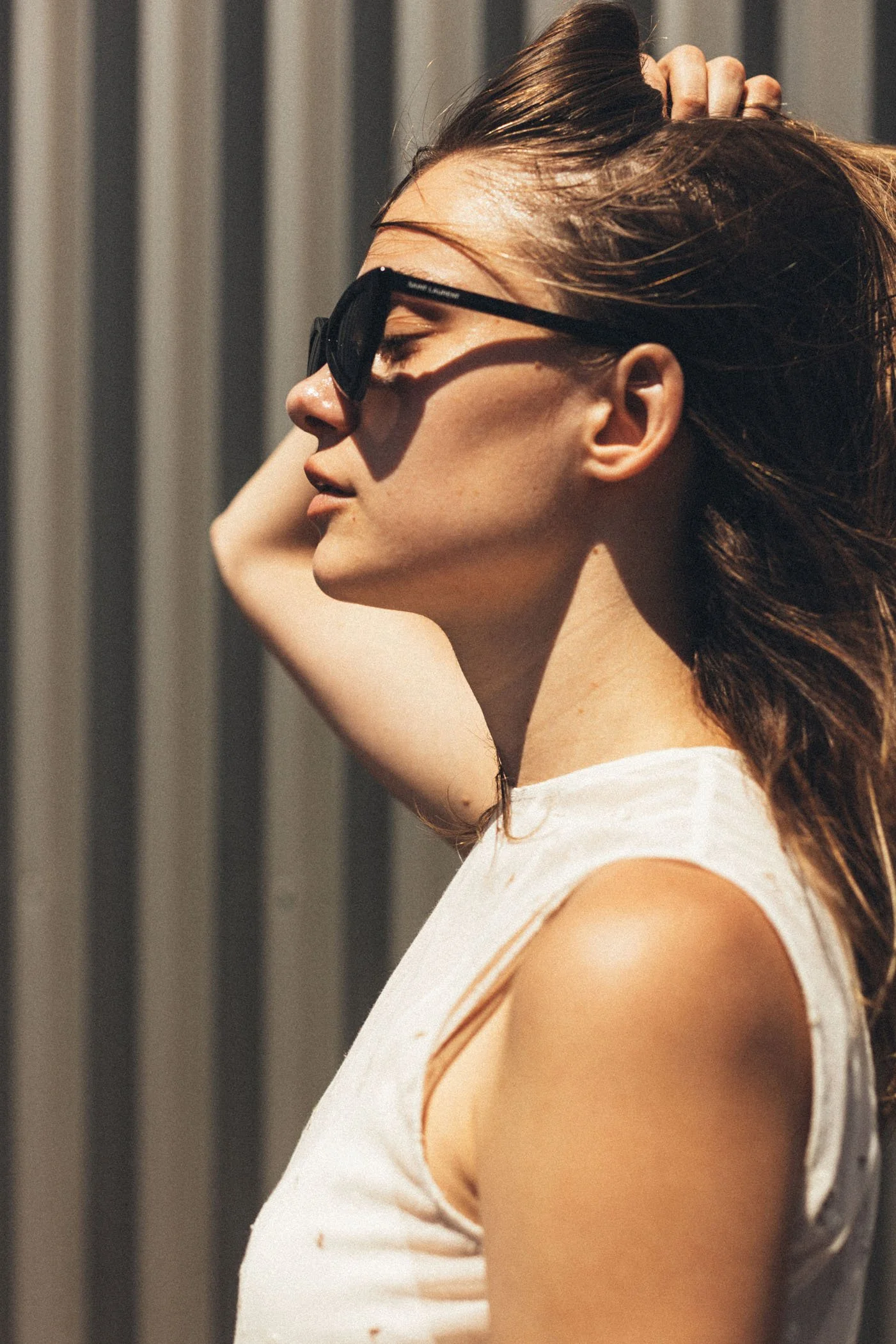 Mujer con gafas oscuras, cabello rubio, en perfil, tocando su cabello, con fondo de rayas verticales.