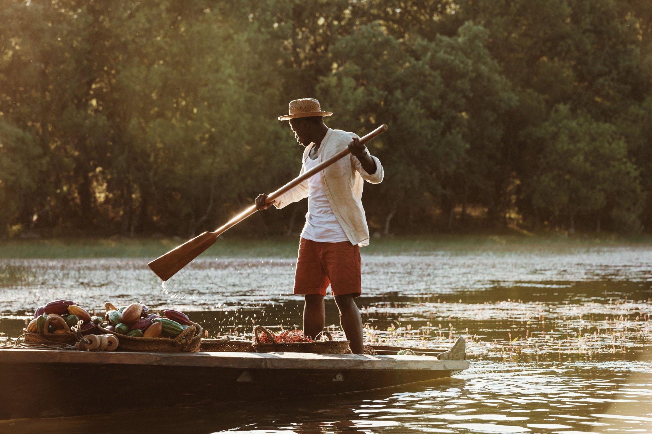 Joven en un barco en el río, remando con un largo bastón, con un sombrero y vista de la naturaleza al atardecer.