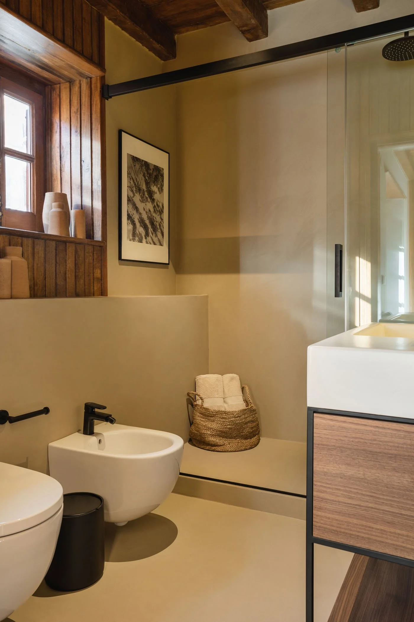 Baño moderno con ducha, bidé, inodoro y lavabo de madera. Hay una ventana con madera, un cuadro en la pared y un cesto de ropa con toallas.