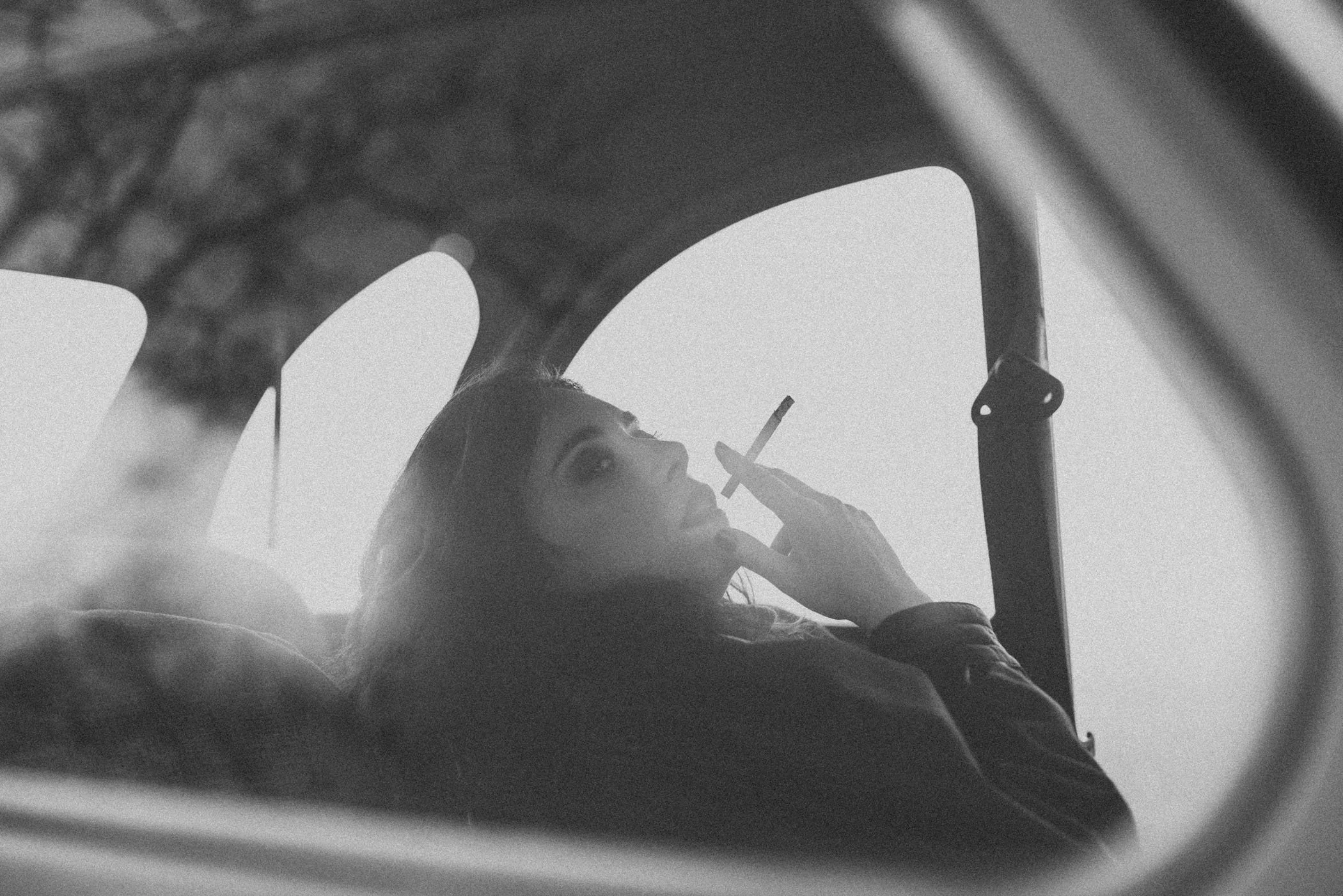 Mujer en coche, vista a través del espejo retrovisor, sosteniendo un cigarrillo, en blanco y negro.
