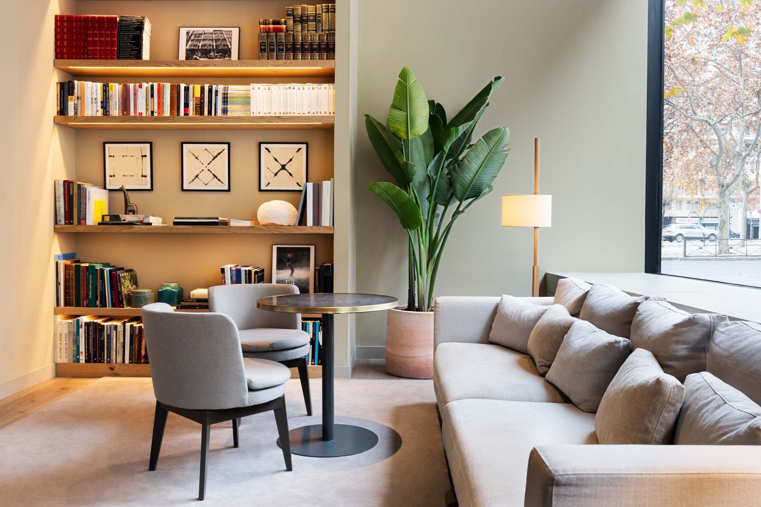 Sala de estar con estantería llena de libros, un sillón, una mesa redonda, un sofá gris, una planta grande en una maceta de color terracota, y una lámpara de pie junto a una ventana con vista a las calles urbanas.