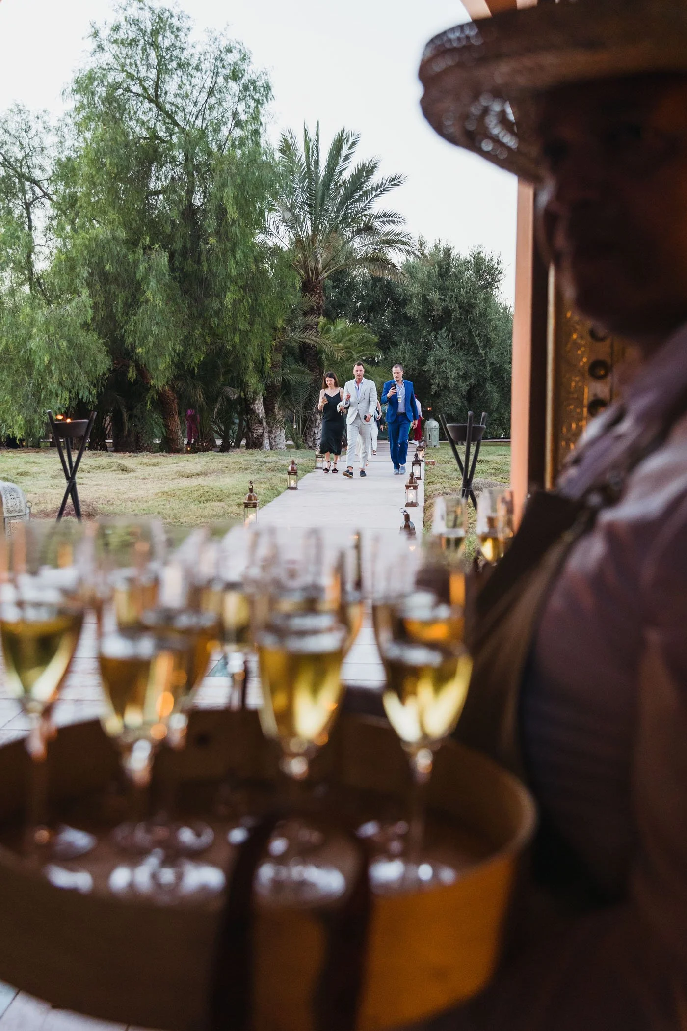 Una persona sostiene una bandeja con varias copas de vino blanco, mientras que en el fondo se ven varias personas caminando por un sendero en un jardín con árboles verdes.