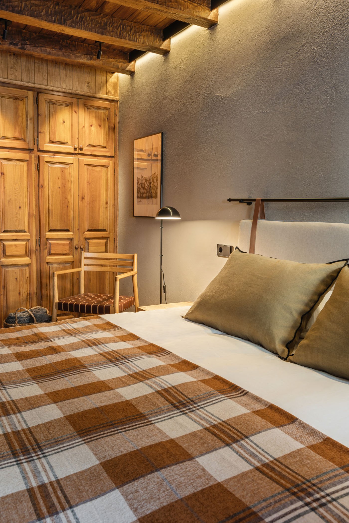 Dormitorio con paredes de color claro, cama con sábanas blancas y almohadas en tonos marrón, manta a cuadros en tonos marrón y beige, armario de madera, silla de madera con cojín a rayas en tonos marrón y beige, lámpara de piso junto a la cama y cuad