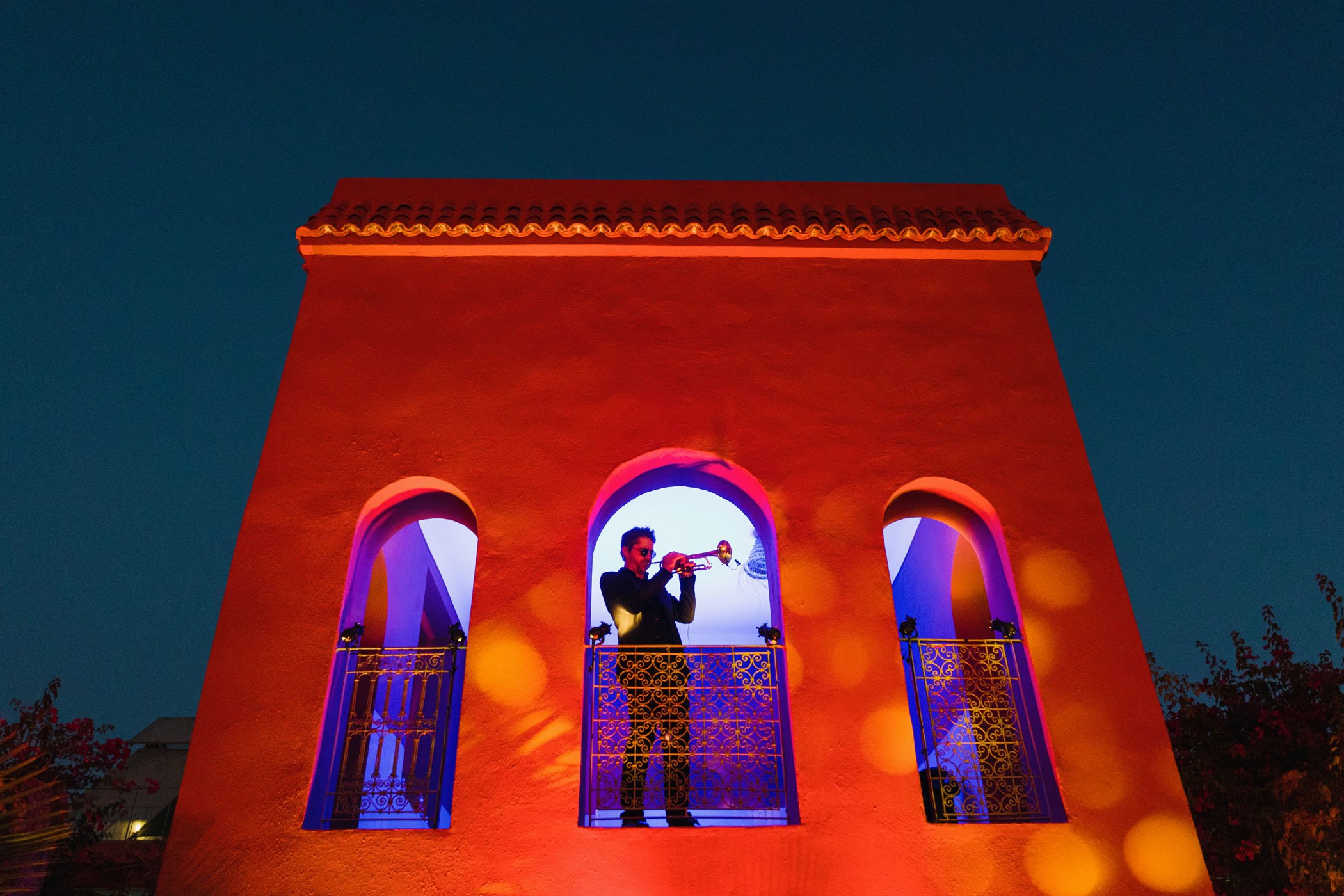 Un músico tocando la trompeta en un balcón de un edificio naranja con arco en la noche, con farolas visibles y un cielo oscuro de fondo.