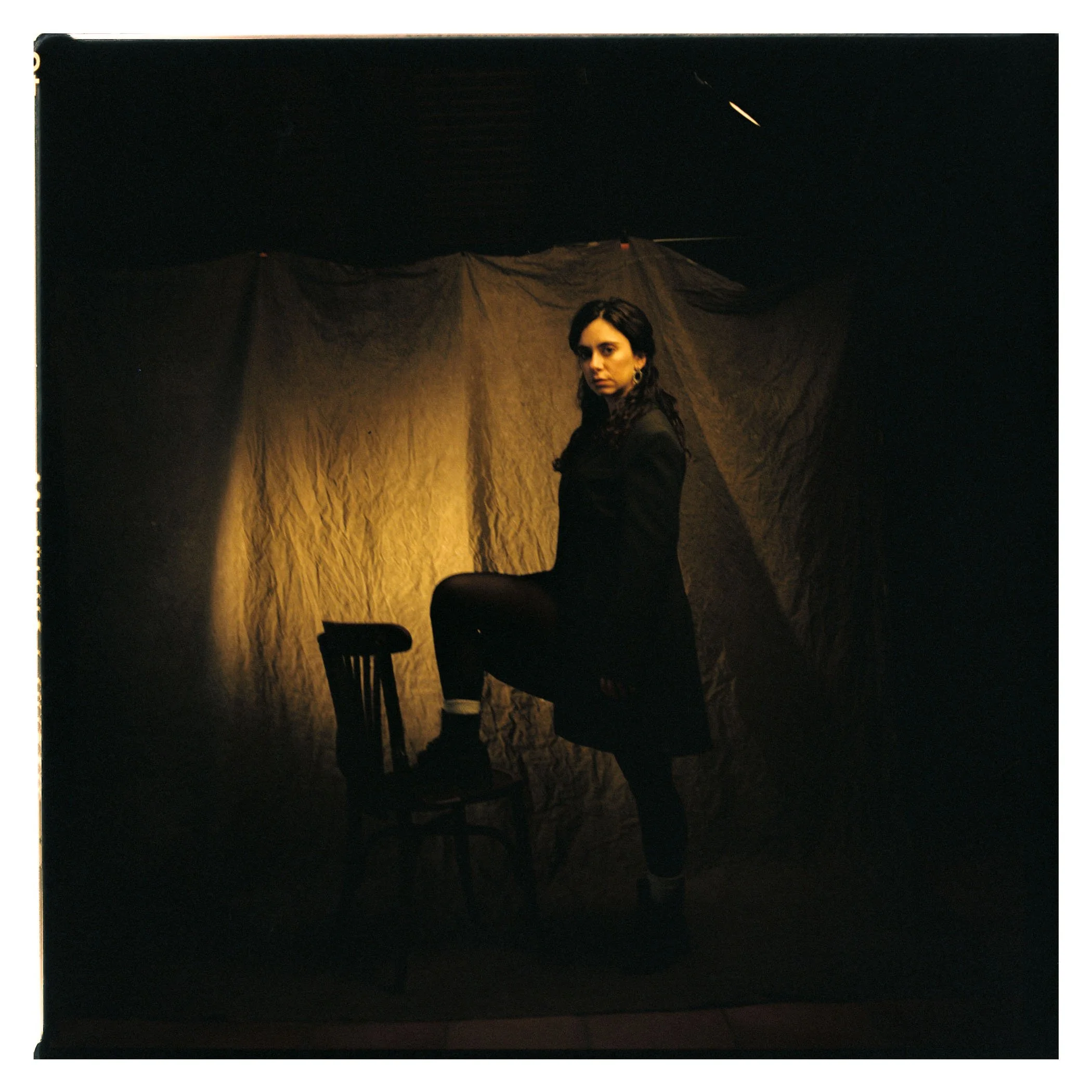 Mujer de pie con una pierna sobre una silla, vestida con ropa oscura, en un fondo oscuro con una luz cálida."}