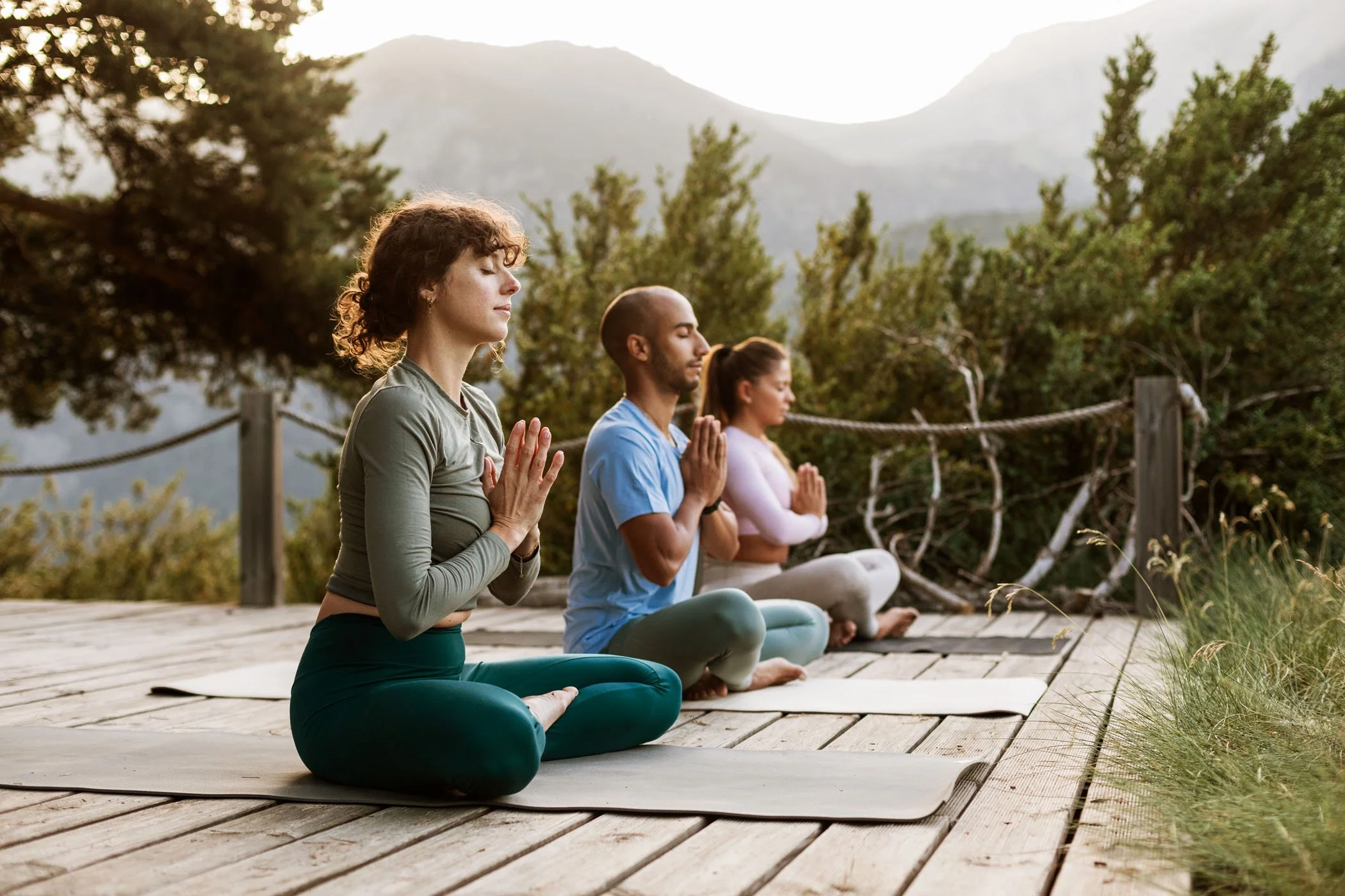 Tres personas practicando yoga en un bote de madera al aire libre, rodeadas de naturaleza y montañas al fondo, en primavera o verano.
