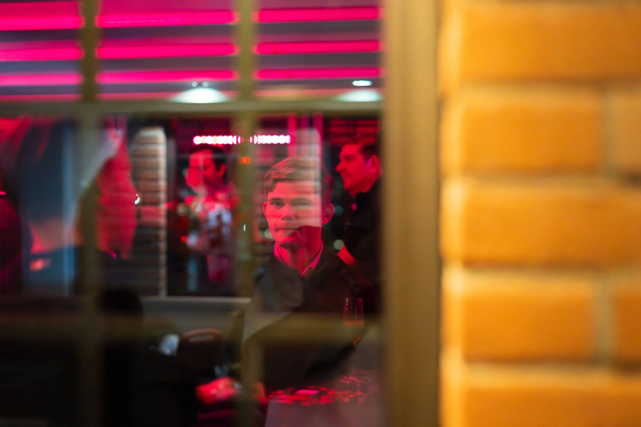 Hombre sentado en un restaurante o bar, visto a través de una ventana con rejas, con luces rojas y fondo de otras personas en un ambiente social.
