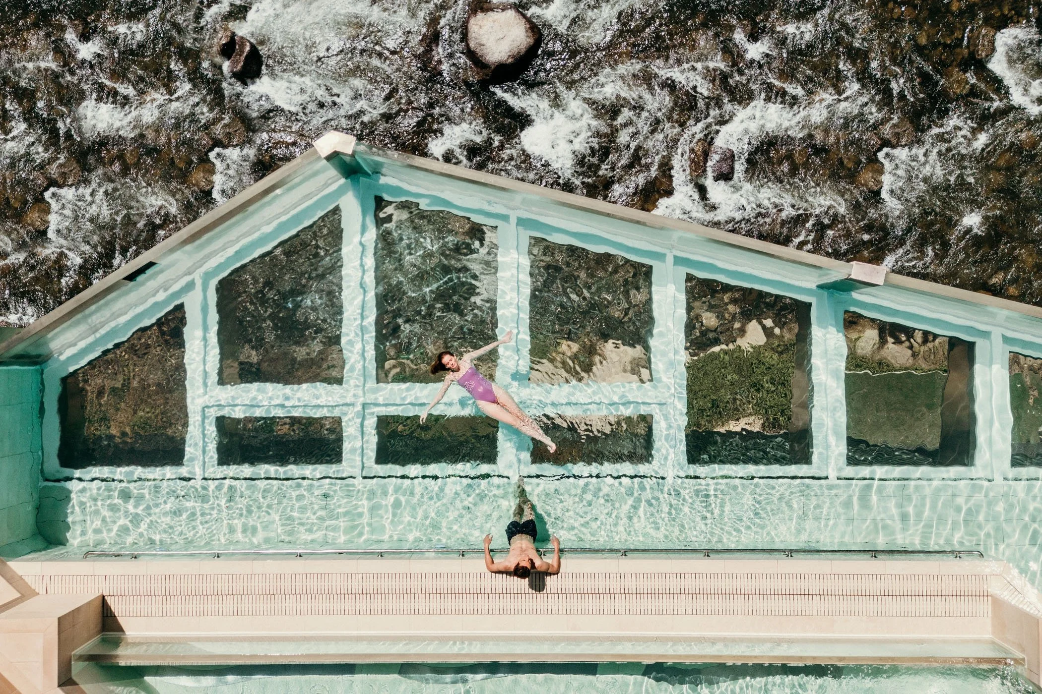 Una piscina de cristal en un edificio alto con dos personas dentro, una de ellas en traje de baño y otra en traje de baño rosa, con vista a un río o río de agua rápida.