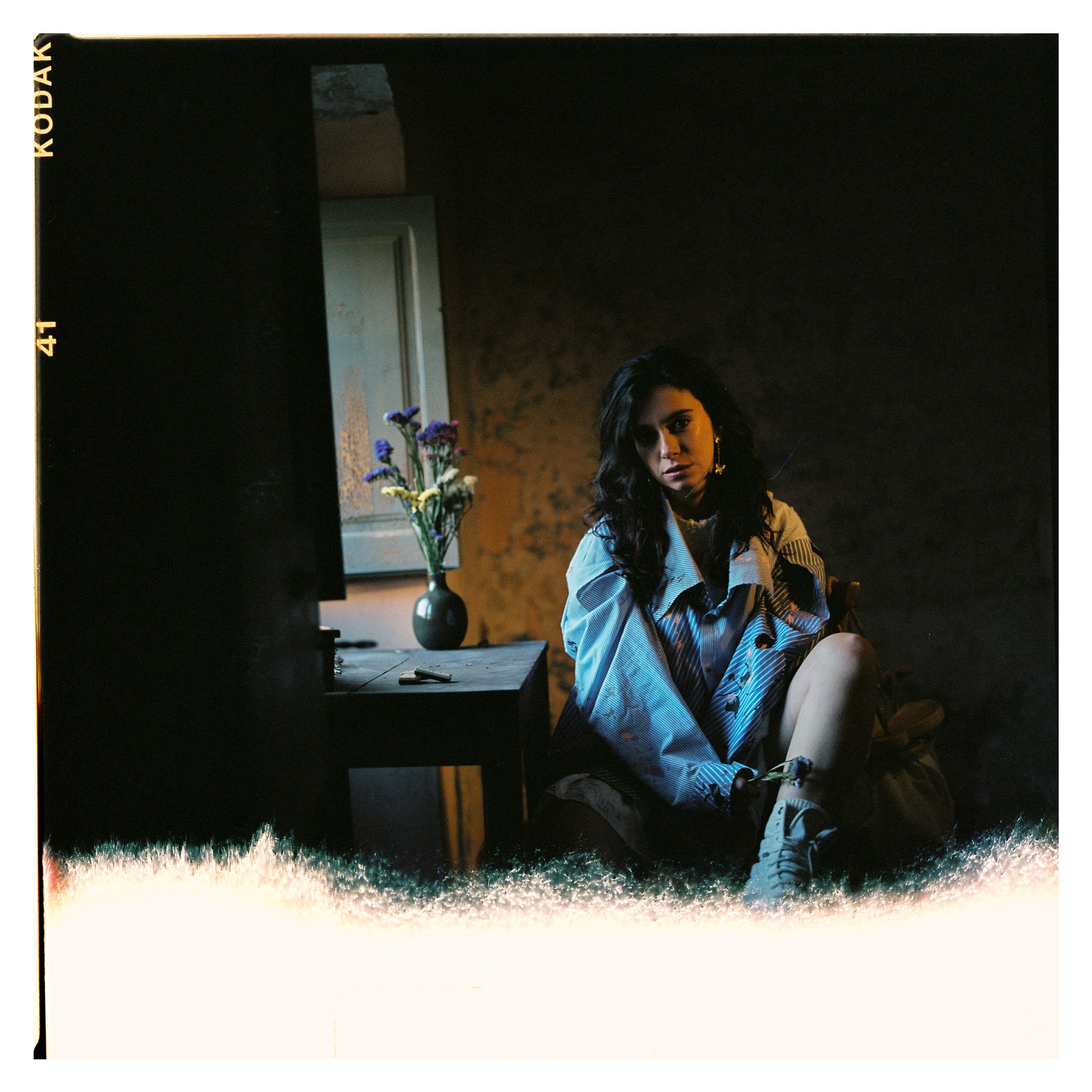 Una mujer con cabello oscuro y despeinado, usando una chaqueta de cuadros, sentada en una habitación oscura, con una mesa a su lado con un jarrón de flores y un espejo en la pared.