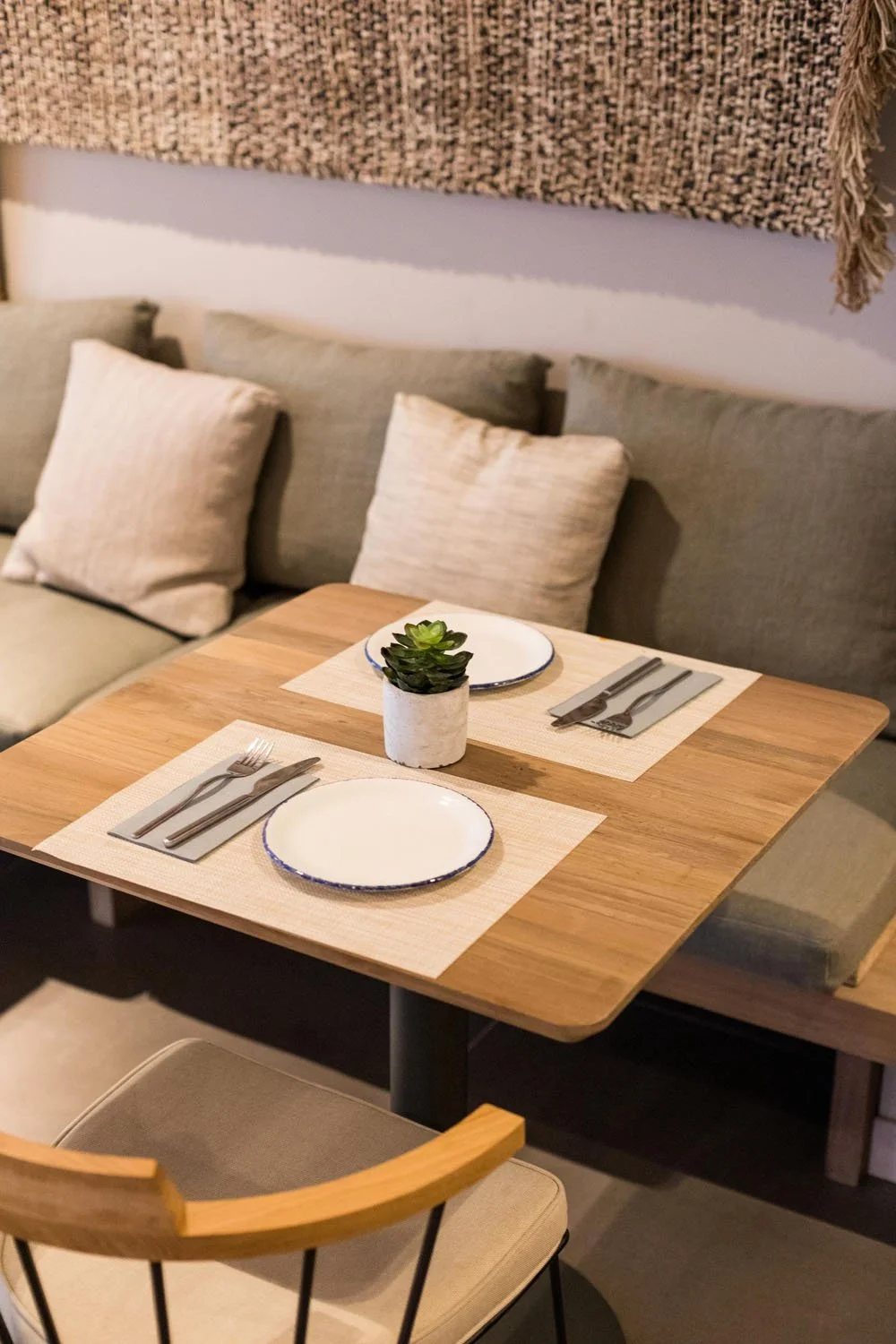 Mesa lista para comer con platos, cubiertos, una planta y un fondo de sofá con cojines en un ambiente acogedor.