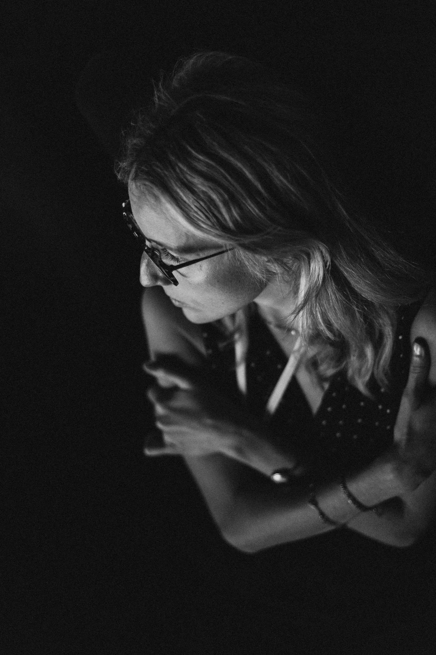 Mujer con gafas y cabello rizado, en expresión pensativa, usando un dispositivo móvil, en un ambiente oscuro.
