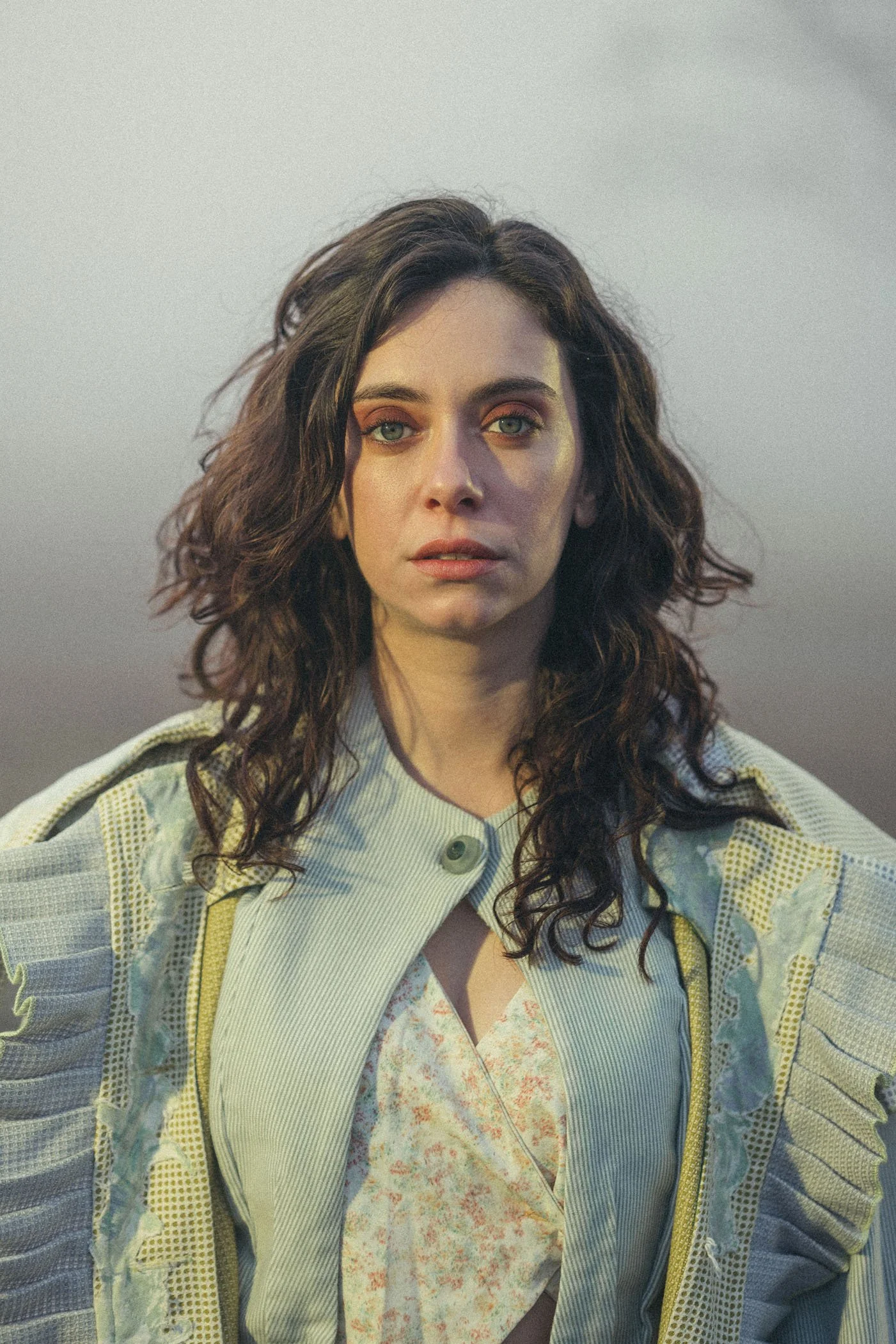 Retrato de una mujer con cabello rizado y mirada intensa, vestida con una chaqueta y blusa en tonos claros, en un fondo gris difuso.
