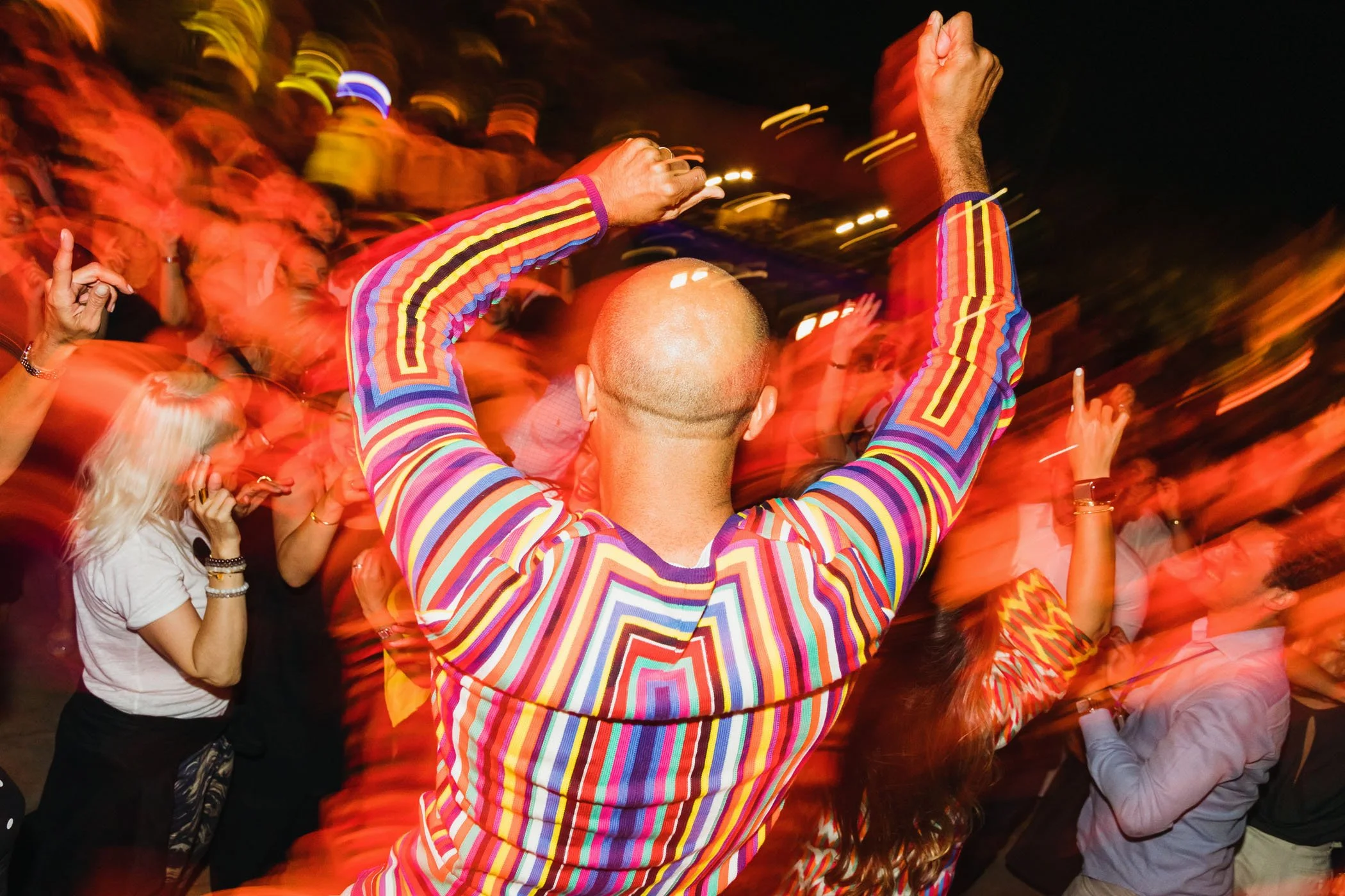 Personas bailando y disfrutando en una fiesta nocturna con movimiento y luces coloridas, destacando a una persona en primer plano con un suéter de rayas multicolores, levantando los brazos.