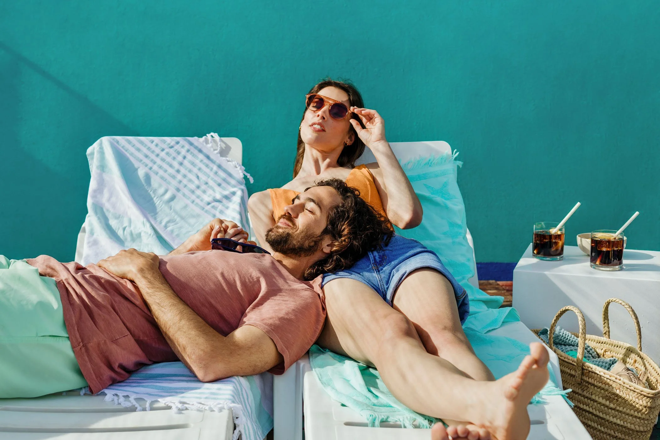 Una pareja tomando el sol en una tumbona junto a una mesa con bebidas. La mujer lleva gafas de sol y una blusa naranja, el hombre tiene barba y lleva ropa de verano. Ambos parecen relajados y disfrutando del día.