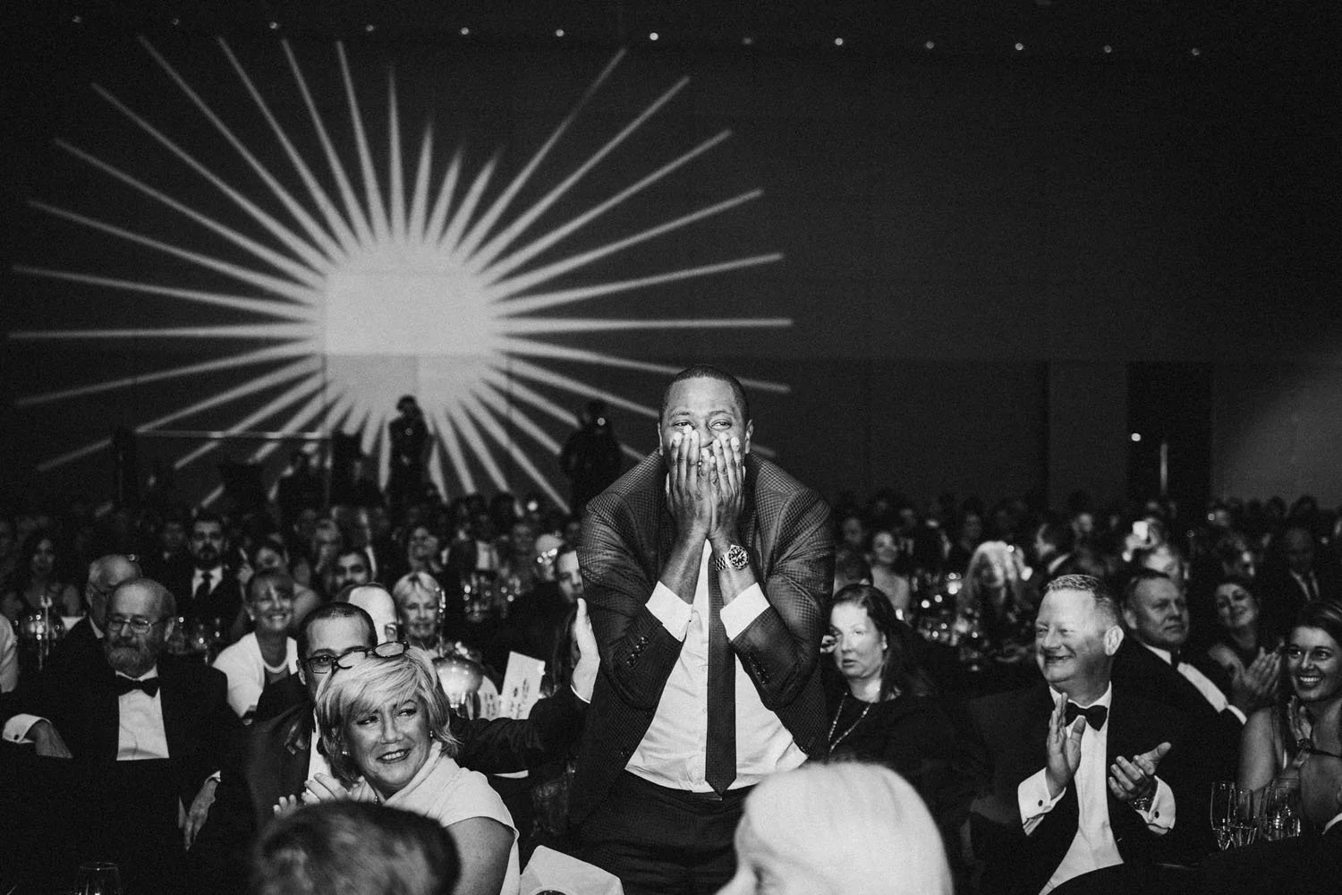 Un hombre en traje de novio con la mano en la cara y sonriendo en una cena de gala, rodeado de otras personas también vestidas formalmente, en un salón con iluminación artística en la pared de fondo.