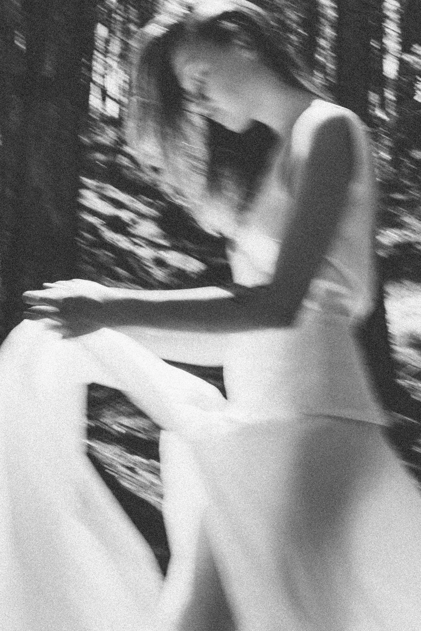 Una mujer con cabello largo, vestida con un vestido largo y blanco, sentada en un entorno boscoso, en una foto en blanco y negro con efecto de movimiento.