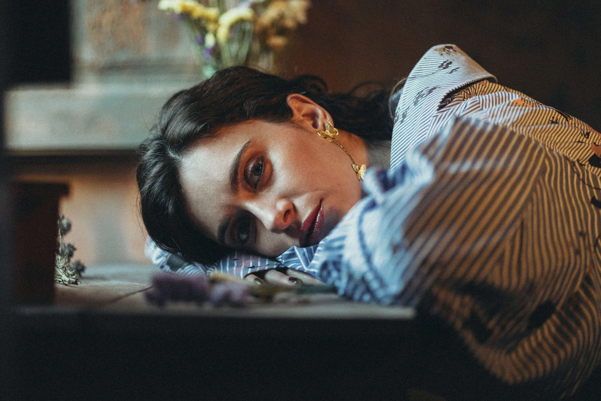 Mujer acostada en una cama, con expresión triste, usando ropa con rayas y accesorios dorados.