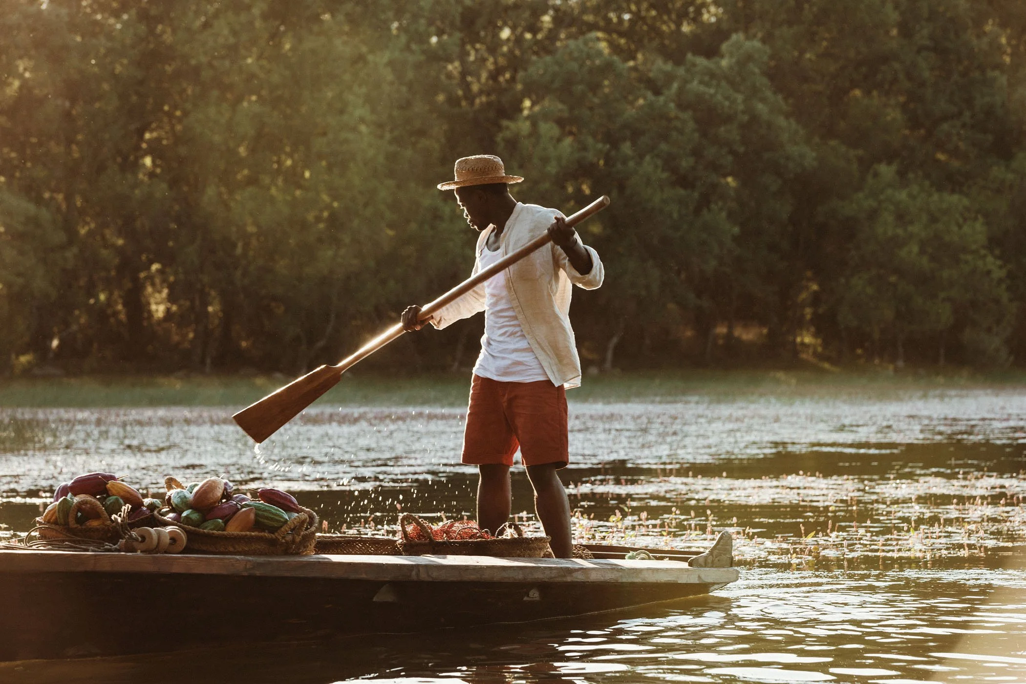 Joven pescador con sombrero usa una pala para pescar en un bote en un río rodeado de árboles en el atardecer.