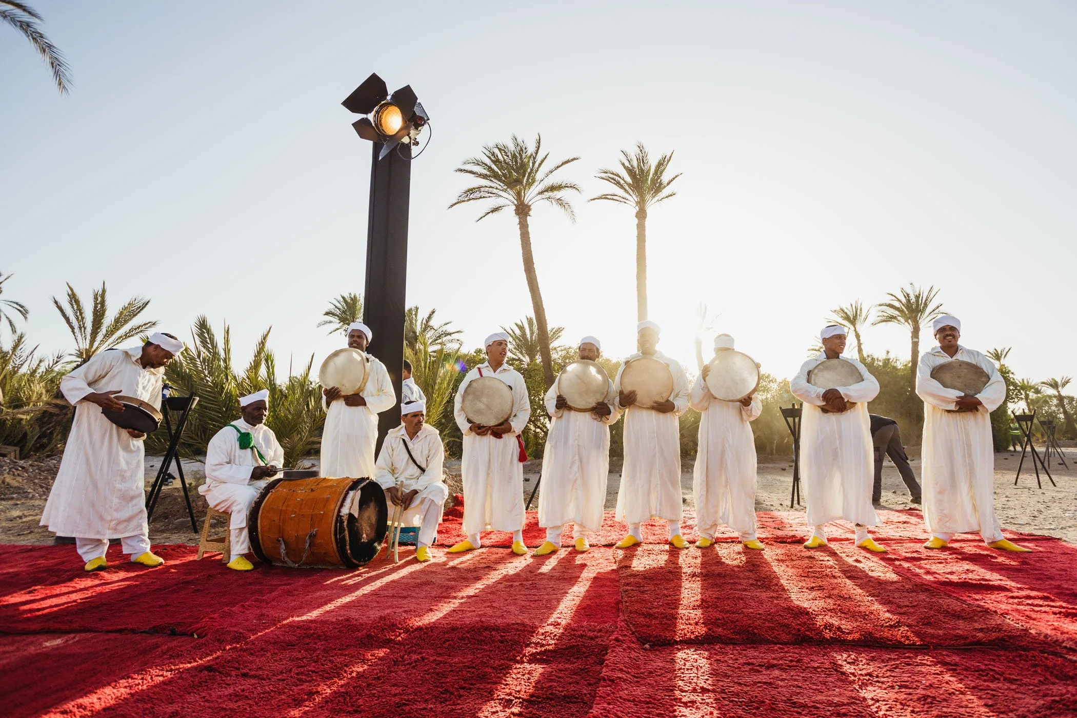 Grupo de músicos tocando instrumentos tradicionales en un escenario al aire libre con palmeras y sol de fondo.