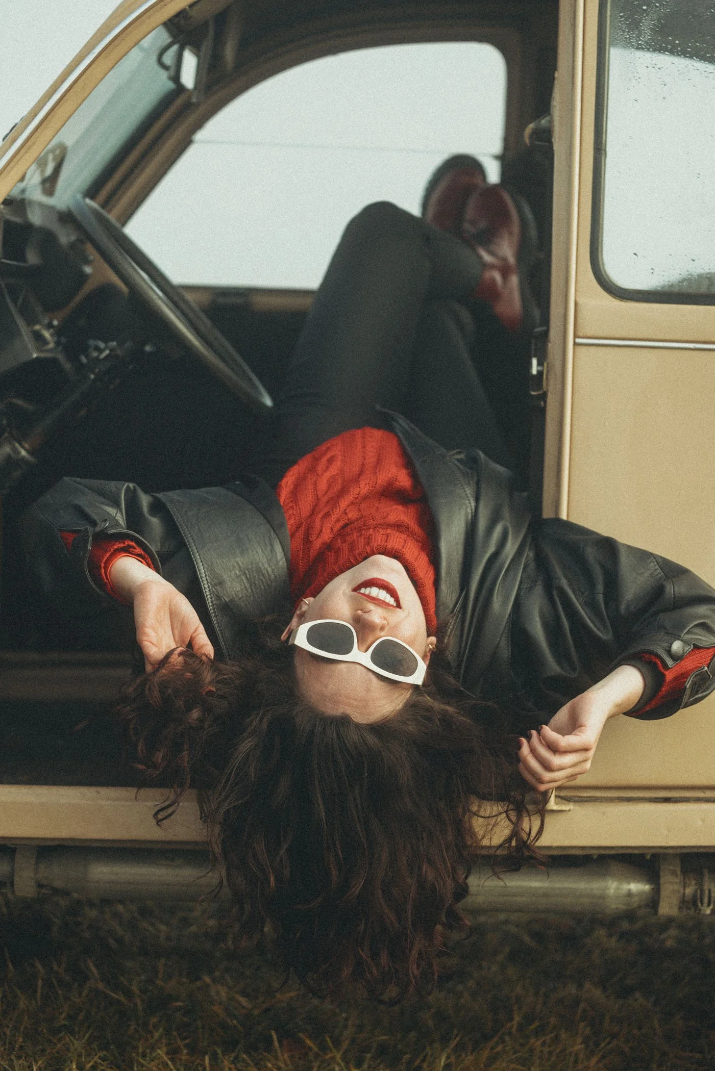 Una mujer acostada en el suelo, con gafas blancas, cabello rizado, pantalones negros, una chaqueta de cuero y un suéter rojo, recargada en la entrada de una furgoneta beige en un ambiente exterior.