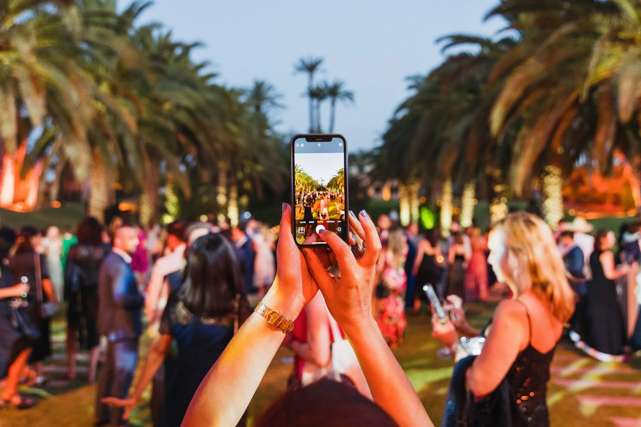 Persona tomando una foto con un teléfono en una fiesta al aire libre con muchas personas y palmeras de fondo.