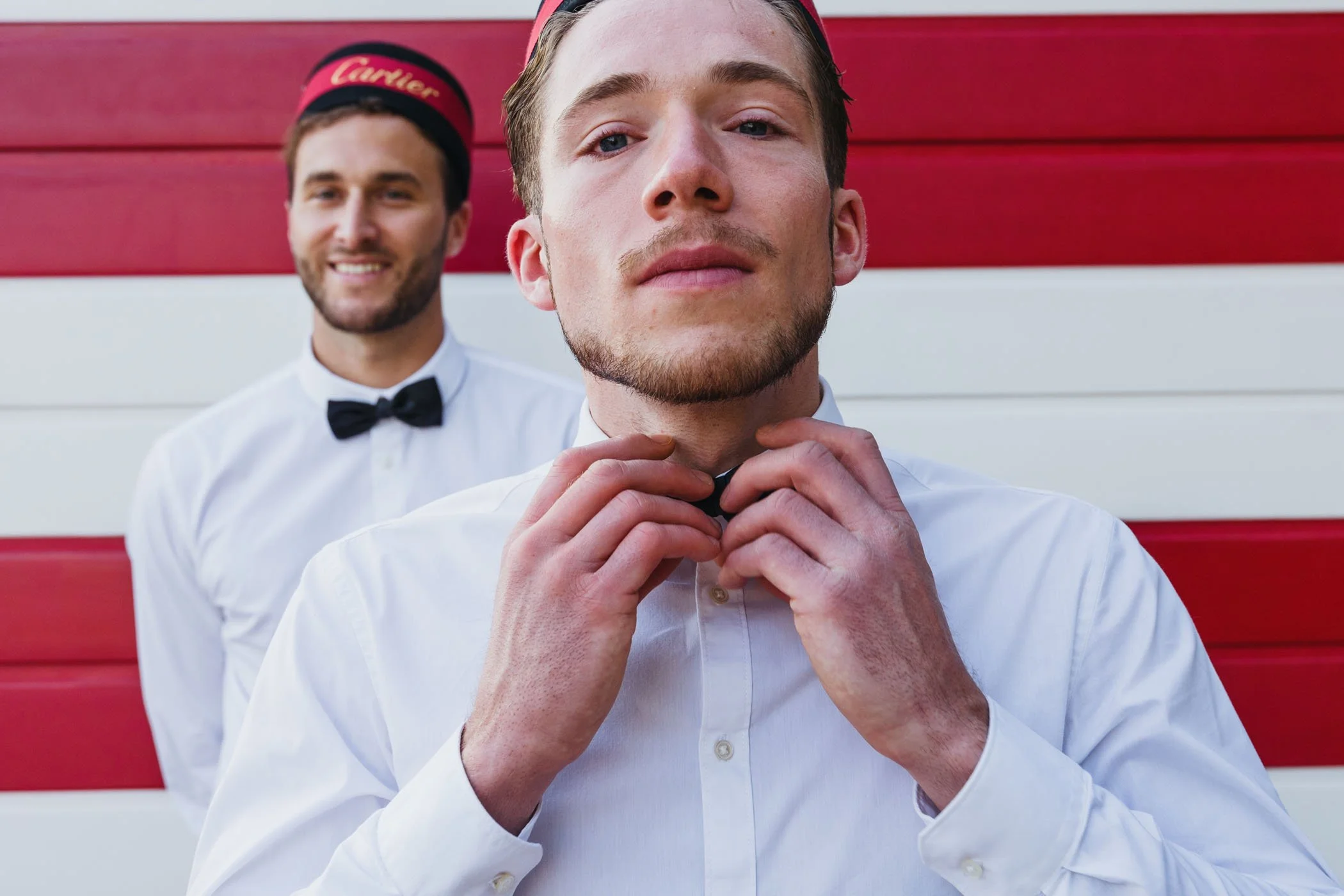 Dos hombres jóvenes en camisa blanca, uno ajustándose el lazo del cuello, el otro en el fondo sonriendo con ropa formal y fondo rojo y blanco.