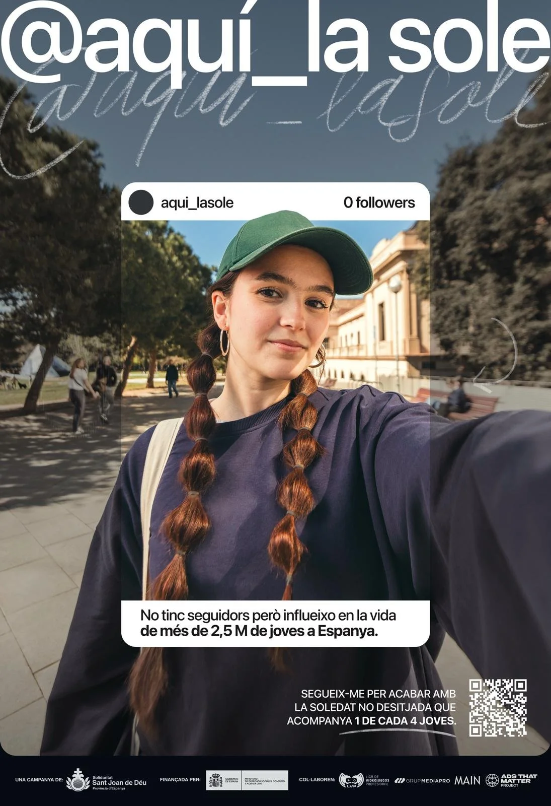 Joven mujer tomando selfie en un parque, con árboles y un edificio antiguo al fondo, usando una gorra verde y camiseta oscura.