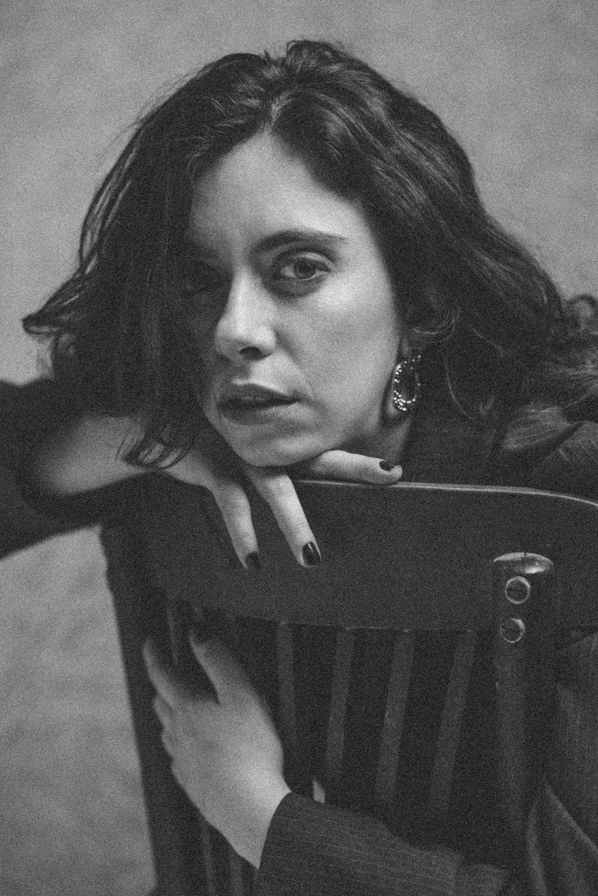 Retrato en blanco y negro de una mujer con cabello rizado, ojos intensos, usando un pendiente grande y un vestido oscuro, apoyada en una silla, mirando directamente a la cámara.
