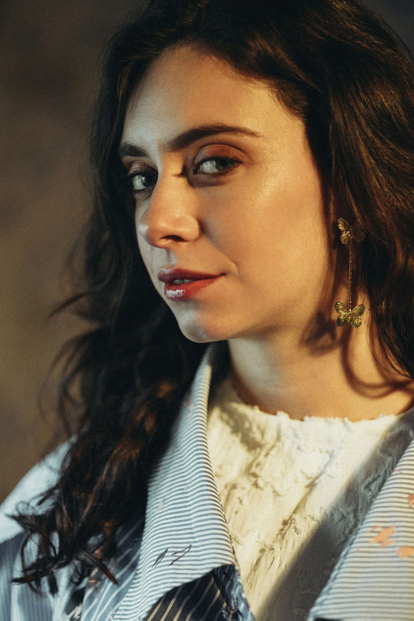 Primer plano de una mujer con cabello castaño oscuro, ojos verdes y labios rojos, usando pendientes de mariposa dorados y un atuendo elegante con una blusa de seda y una chaqueta de rayas.