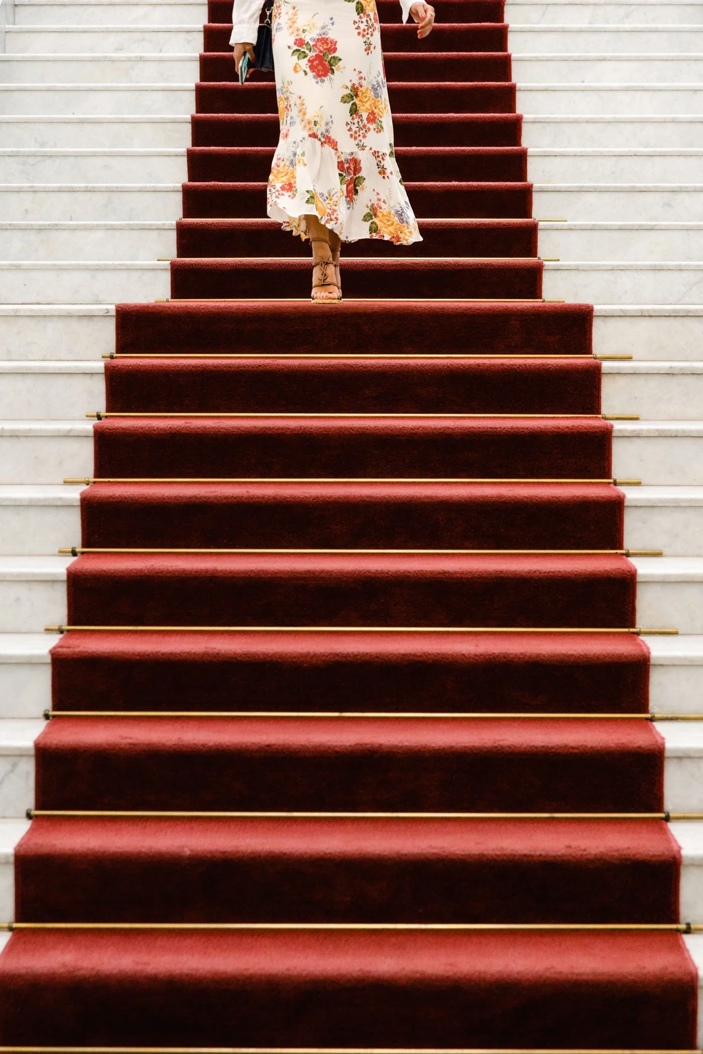 Persona bajando las escaleras cubiertas de alfombra roja, vestida con un vestido largo de flores y tacones.