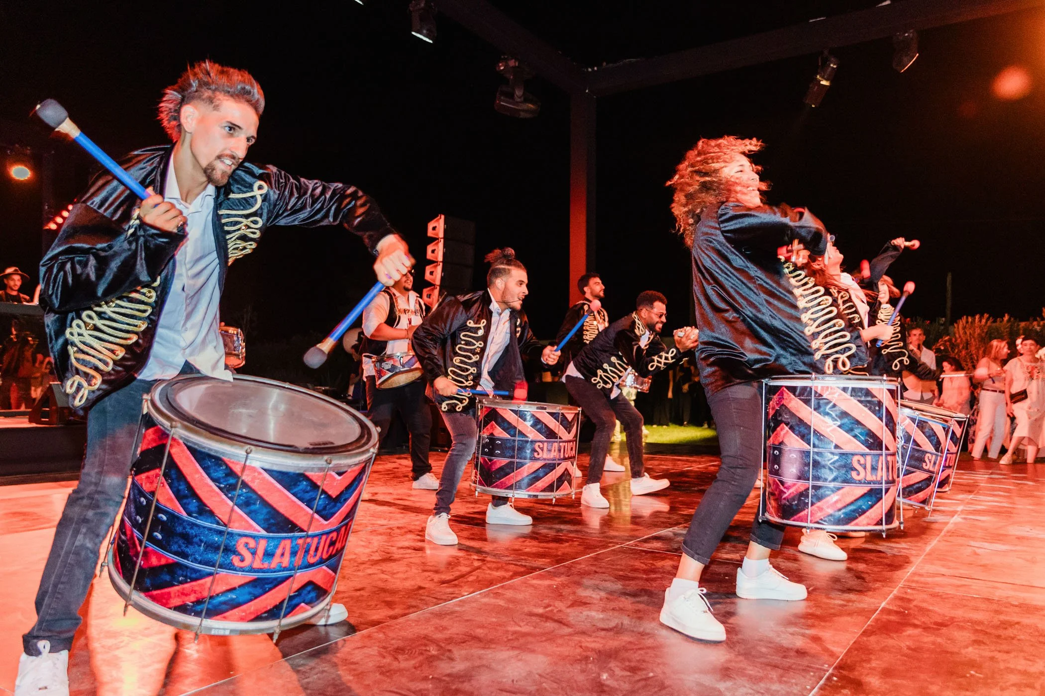 Grupo de personas tocando tambores en un escenario durante una actuación nocturna con ilumincación artística.