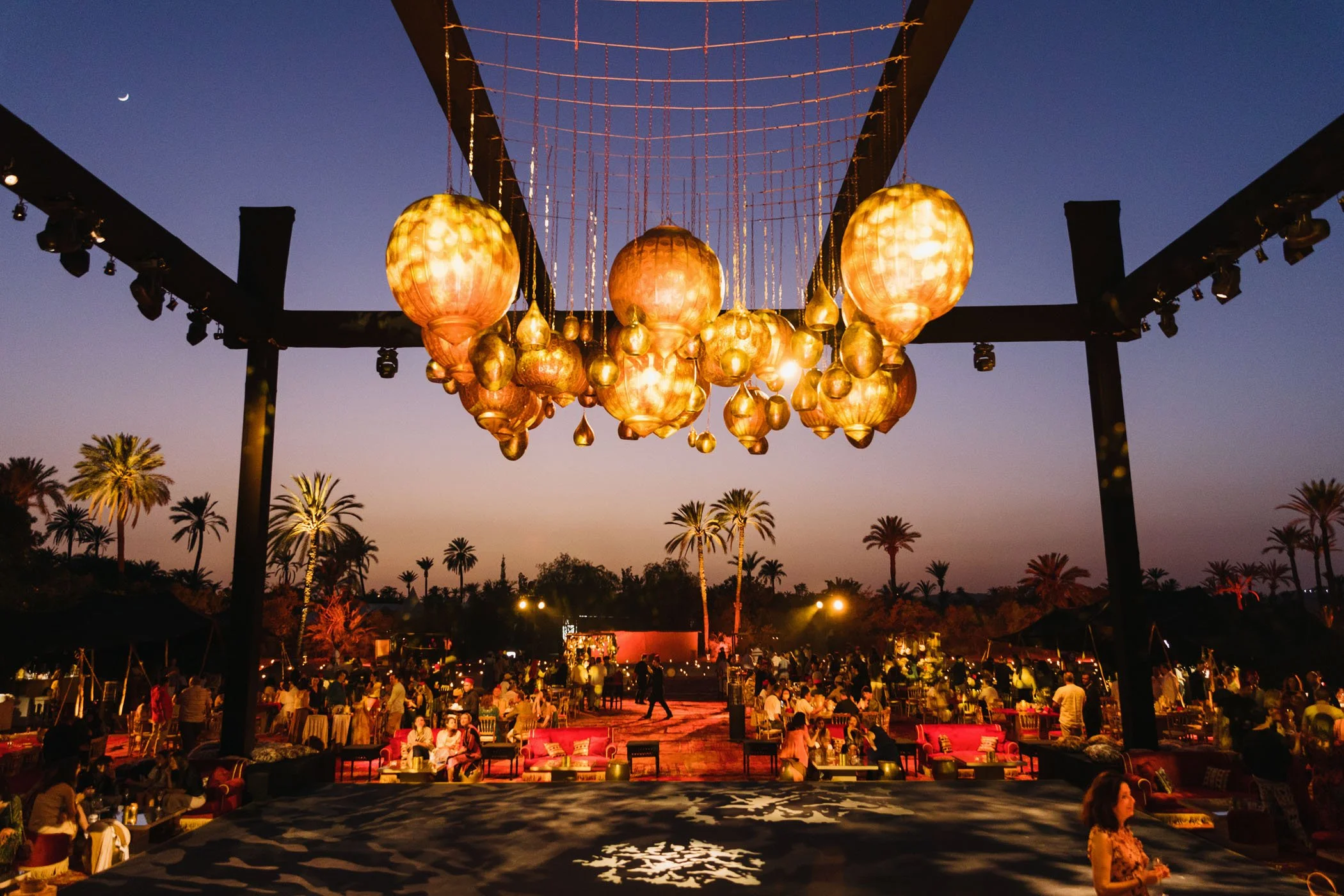 Una vista desde un escenario en un evento al aire libre durante el atardecer, con lámparas decorativas colgantes de color dorado y mesas con personas sentadas, rodeadas de palmeras.