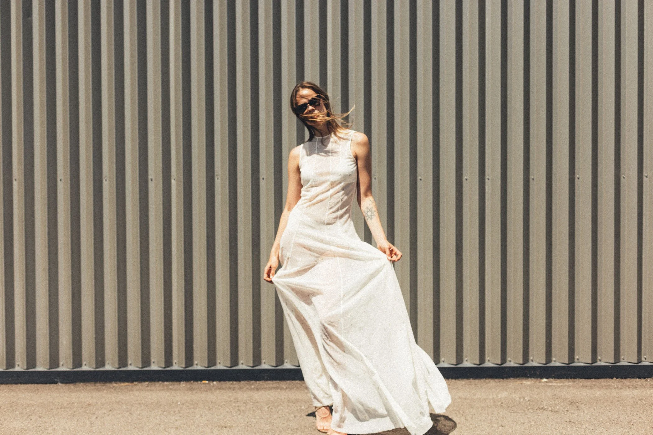 Mujer con vestido largo blanco posando frente a una pared de láminas metálicas enfocada desde abajo.