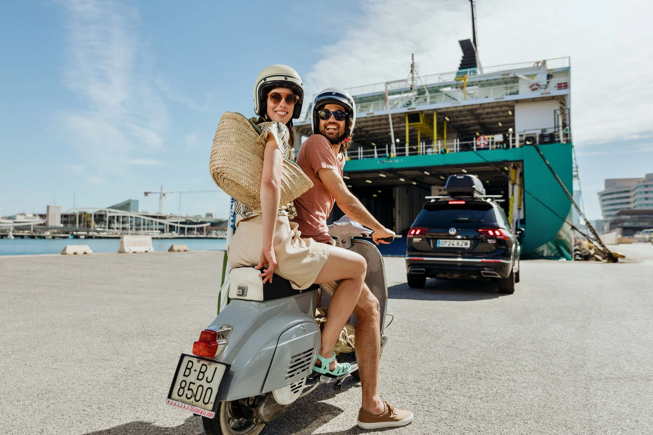 Dos personas con cascos y gafas de sol en una scooter en un muelle, con un barco grande y un coche detrás y edificios en el fondo