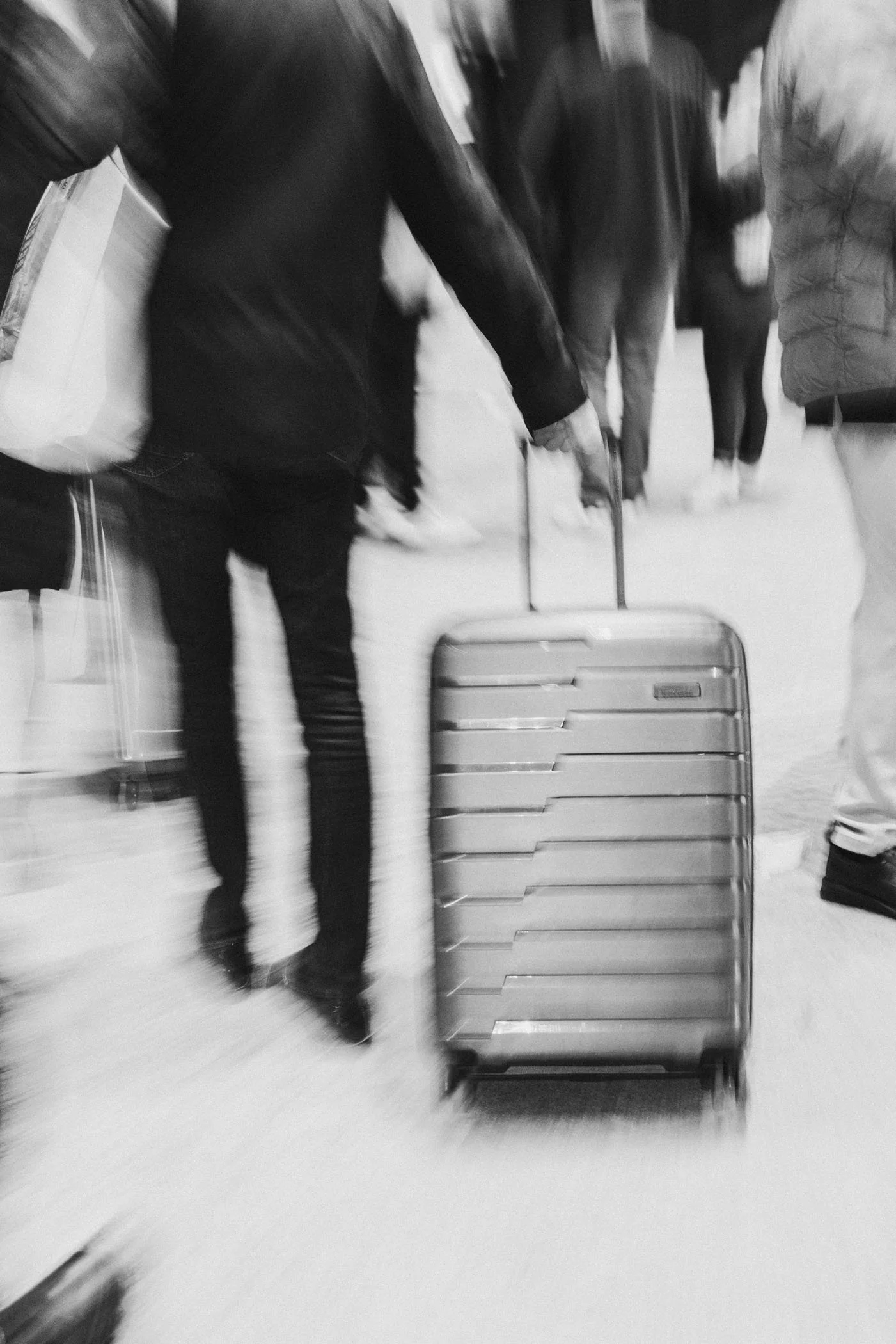 Persona con equipaje de mano en un aeropuerto, caminando entre otras personas. La imagen está en blanco y negro, y muestra movimiento.