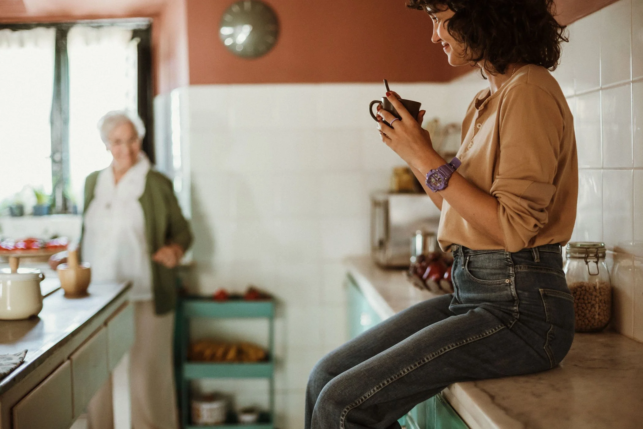 Una mujer joven sentada en la encimera de una cocina, sosteniendo una taza y sonriendo; una mujer mayor de pie en el fondo, cerca de la ventana, con una sonrisa, en una cocina luminosa