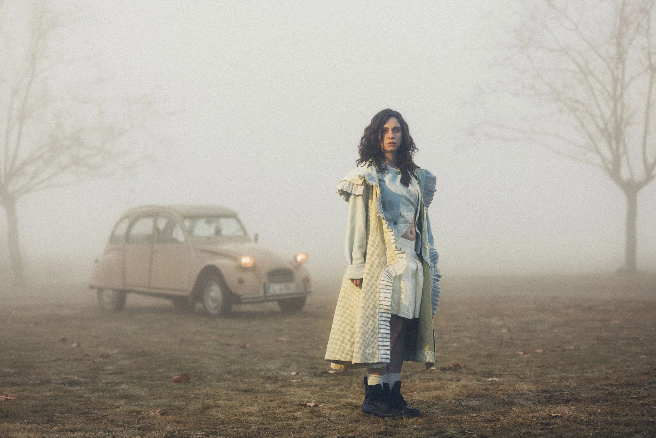 Una mujer de cabello rizado y oscuro está de pie en un campo con niebla, vestida con ropa de colores claros y oscuros. Detrás de ella, hay un auto antiguo y dos árboles sin hojas en la neblina.