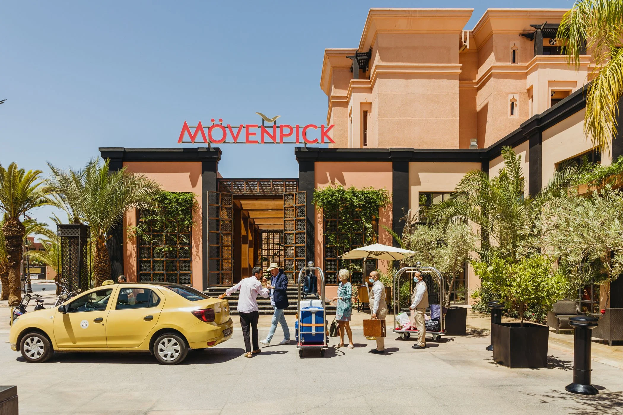 Entrada del hotel Movenpick con personas con equipaje y un coche amarillo frente a la entrada en un día soleado.