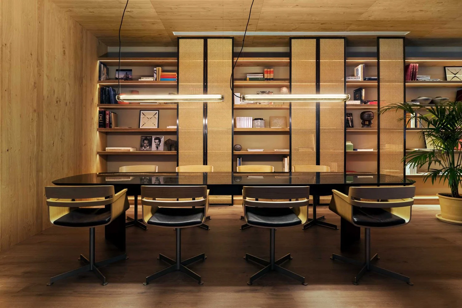 Sala de reuniones con mesa negra, seis sillas de madera y cuero blanco, estanterías con libros y decoraciones, lámparas colgantes y planta en esquina derecha.