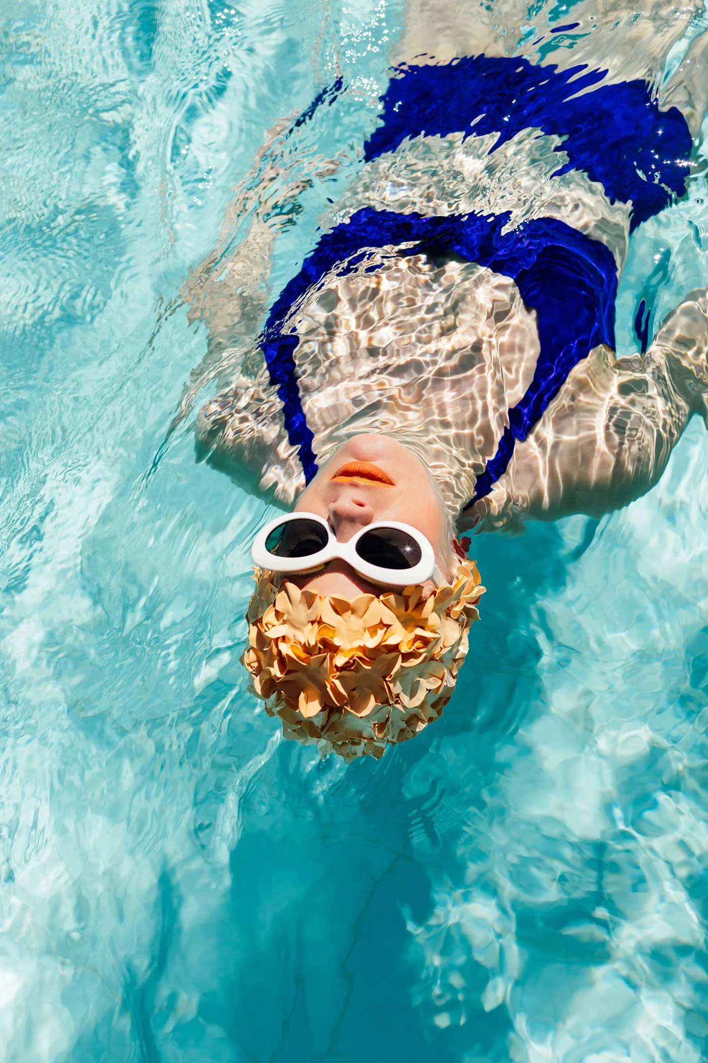 Persona descansando en una piscina, usando gafas de sol grandes, un gorro decorativo y un traje de baño azul, en un día soleado.
